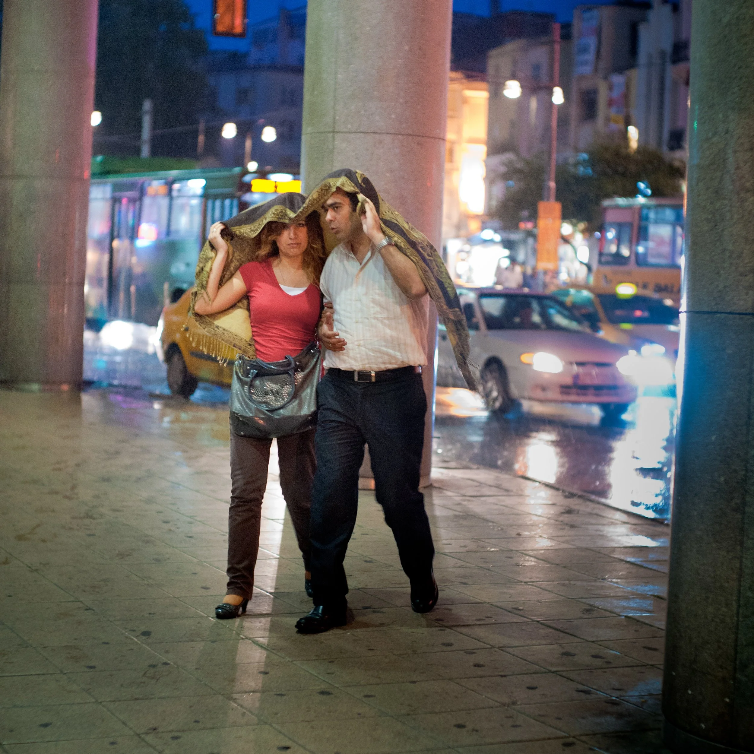 A man and a woman walk under a large piece of cloth shielding them from rain on a city street at night, with traffic and colorful lights in the background.