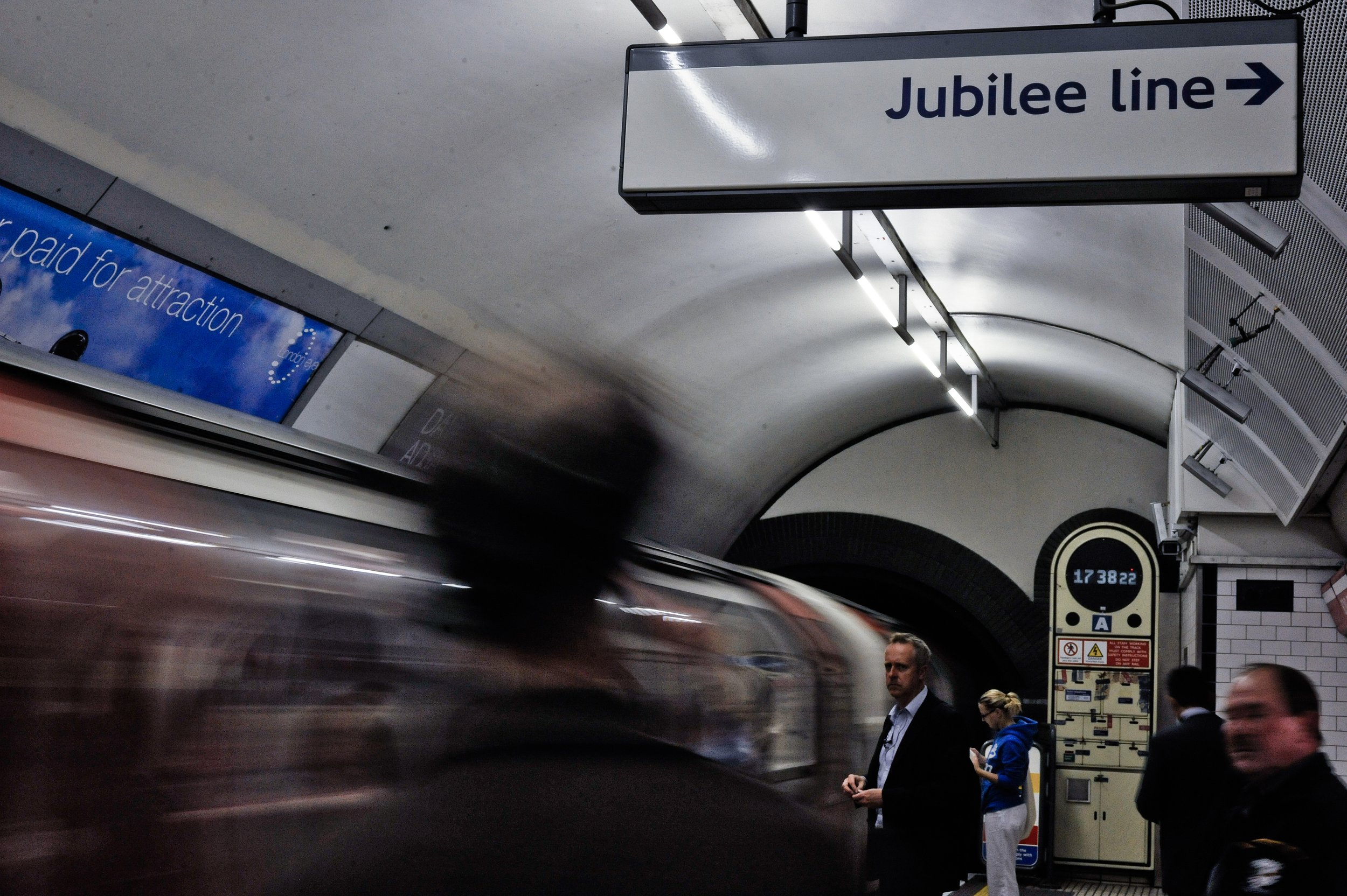 People waiting in the Jubilee line subway station with a moving train passing by. Sign indicating the Jubilee line entrance.