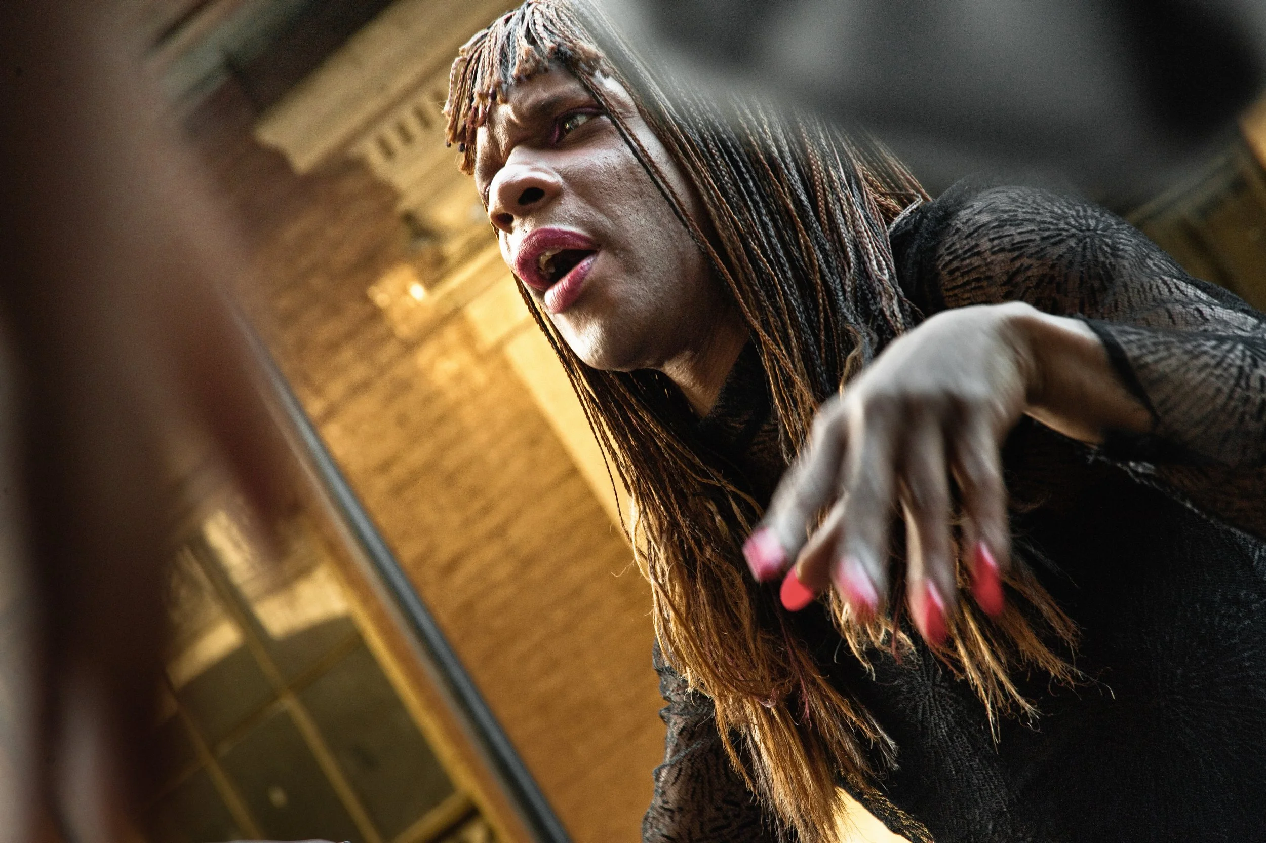 A woman with long, braided hair and pink nails reaches out as she speaks, with a serious expression, indoors with warm lighting.