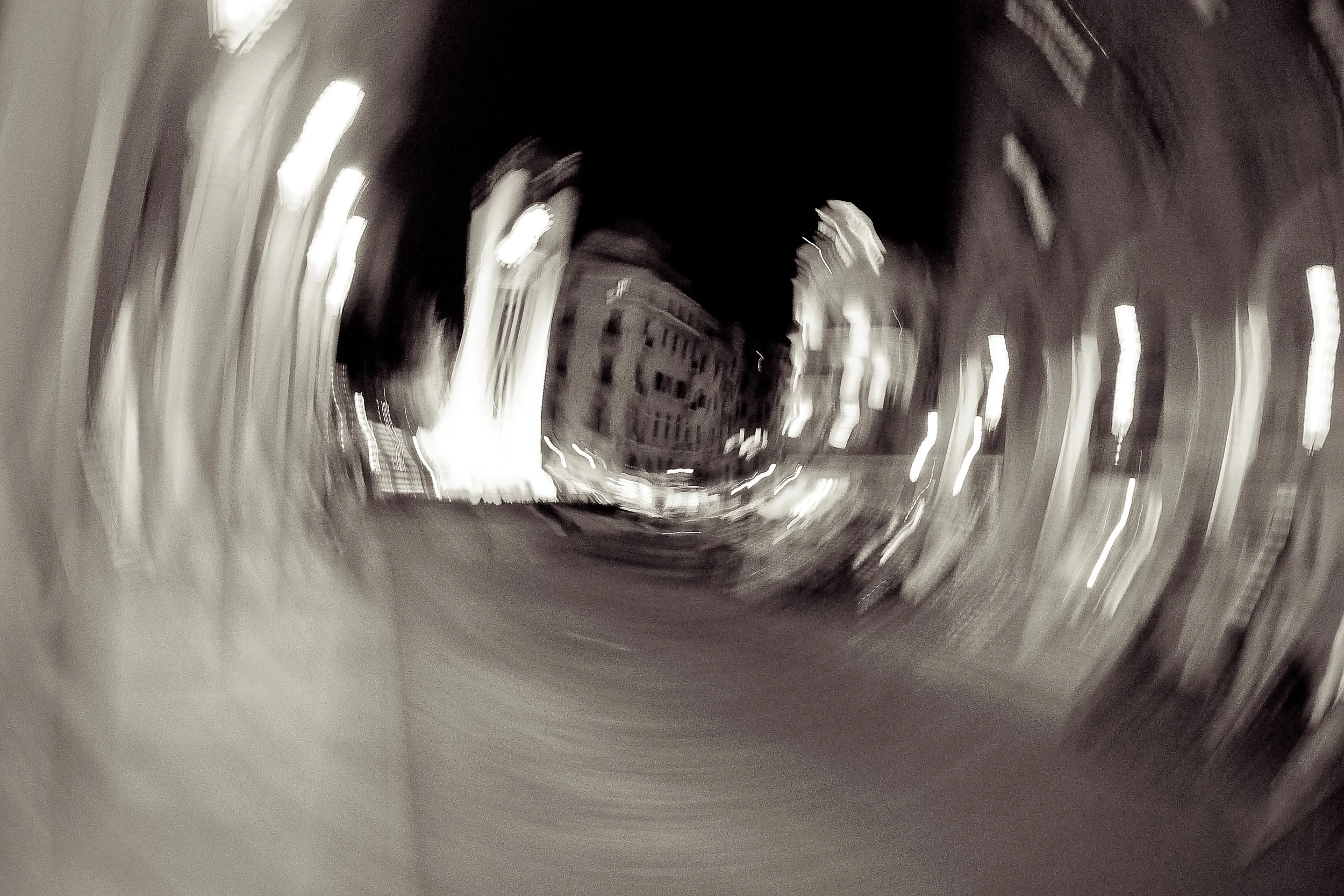 Black and white photo of a city street at night, captured with a motion blur effect creating a swirling, vortex-like appearance.