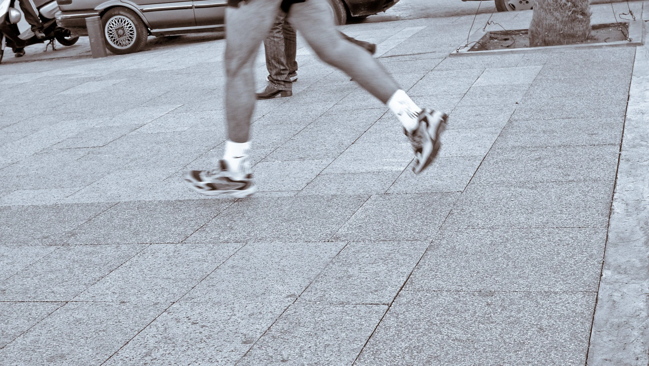 View of a person running on a city sidewalk, focusing on their legs and feet. The person is wearing athletic shoes, white socks, and shorts. In the background, there are parked cars and a tree.