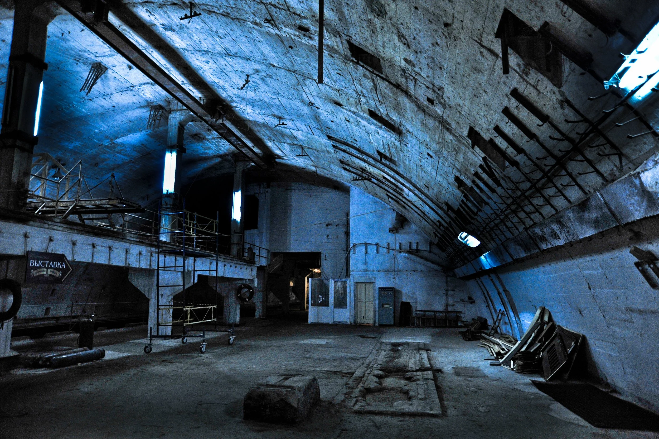 Interior of an empty underground tunnel with a curved ceiling, blue lighting, construction materials, and equipment scattered around.