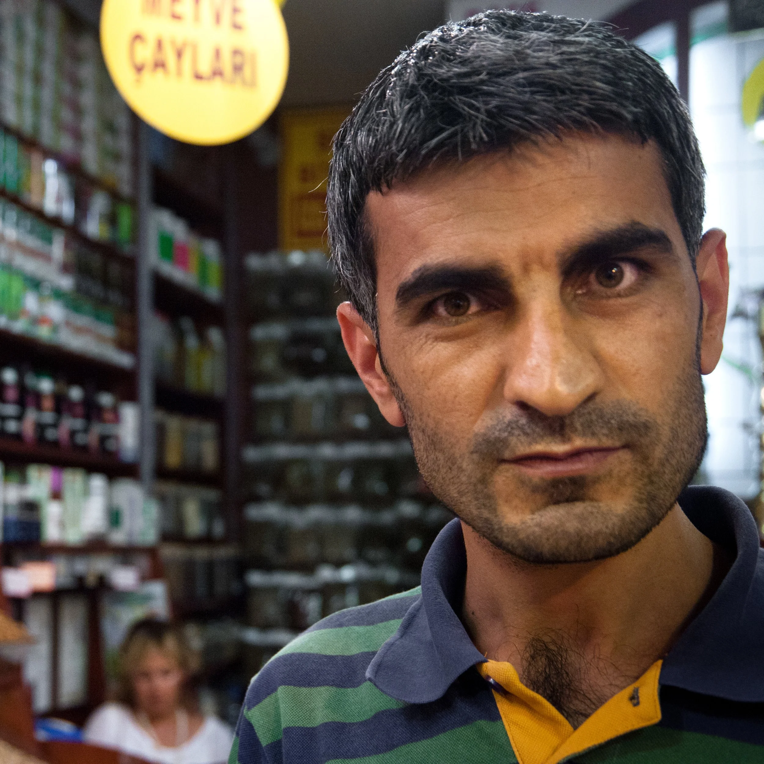 Close-up of a man with dark hair and a striped polo shirt inside a store, with shelves of goods in the background and a woman sitting in the background.