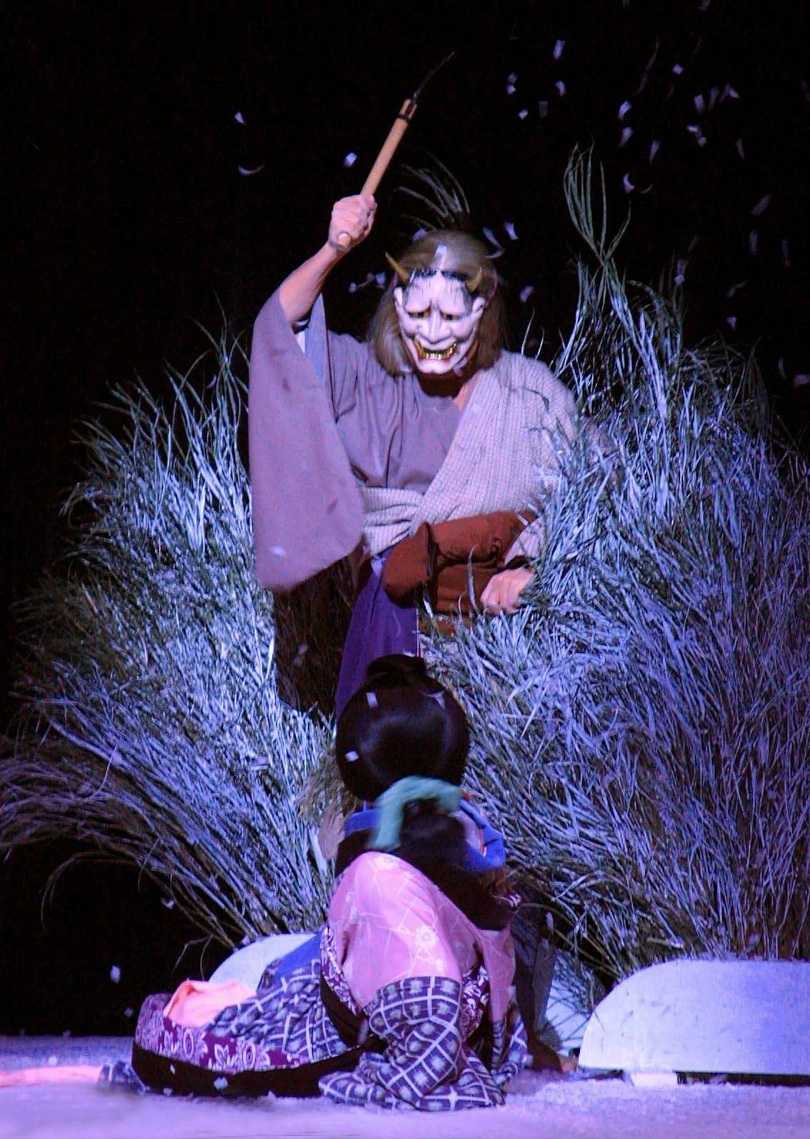 A person dressed as a demon or oni with a horned mask, wielding a stick, standing behind a seated child dressed in traditional Japanese clothing, in front of tall grass with confetti falling around.