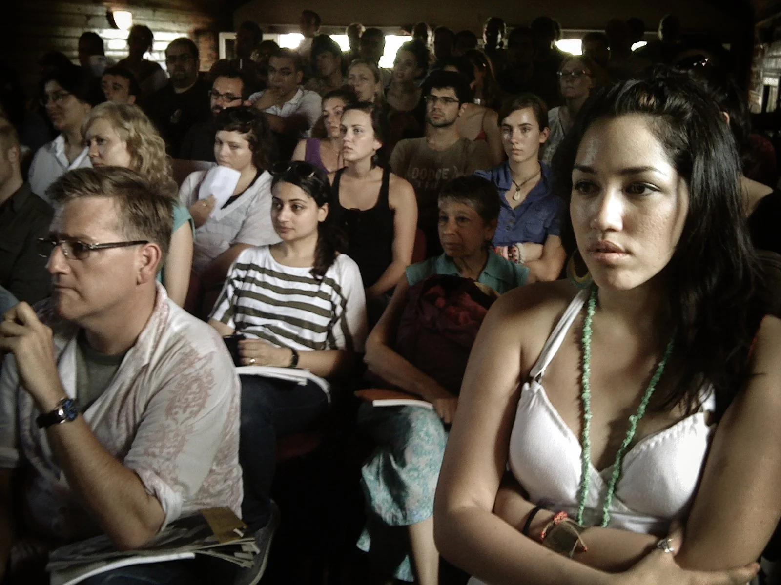 A crowded group of people sitting inside a dimly lit room, attentively listening to an event or presentation.