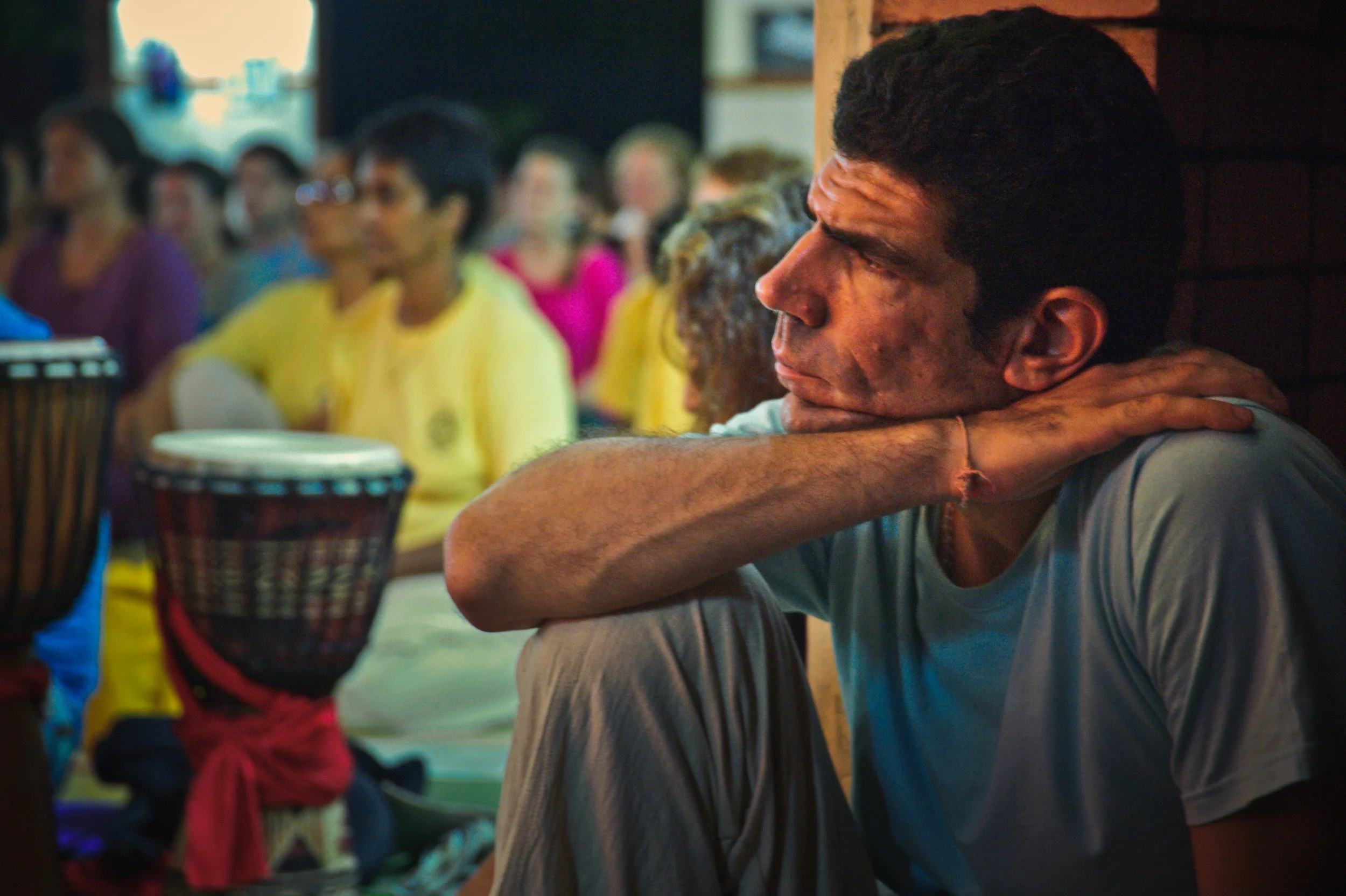 A man sitting with his arm resting on his shoulder, looking contemplative, in a crowded indoor setting with people in colorful clothing in the background.