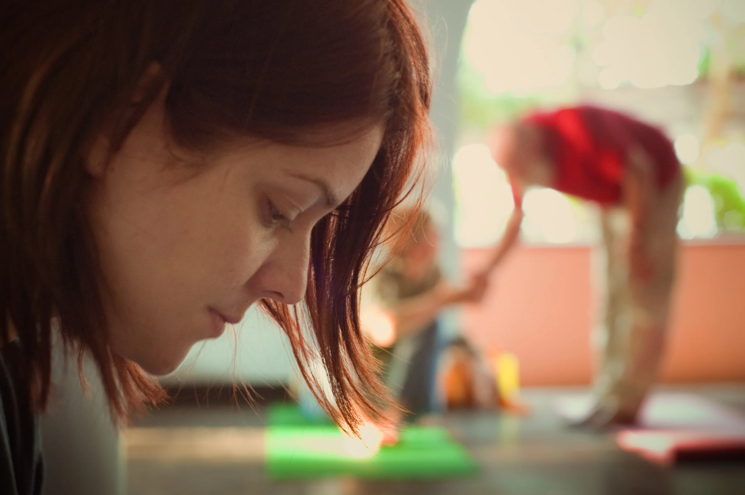 A close-up of a woman with red hair looking down, surrounded by the blurred background of a person with bright red hair and a large dinosaur figure.