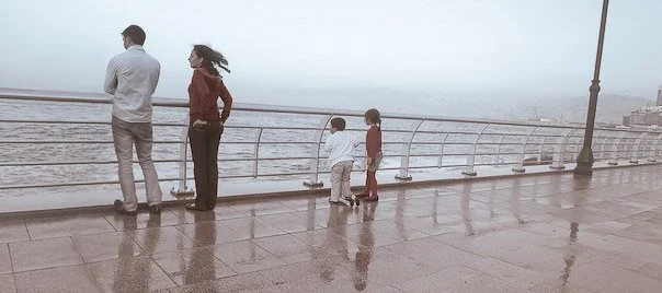 A family of two adults and two children standing on a wet pier, looking out at the ocean on a foggy day.