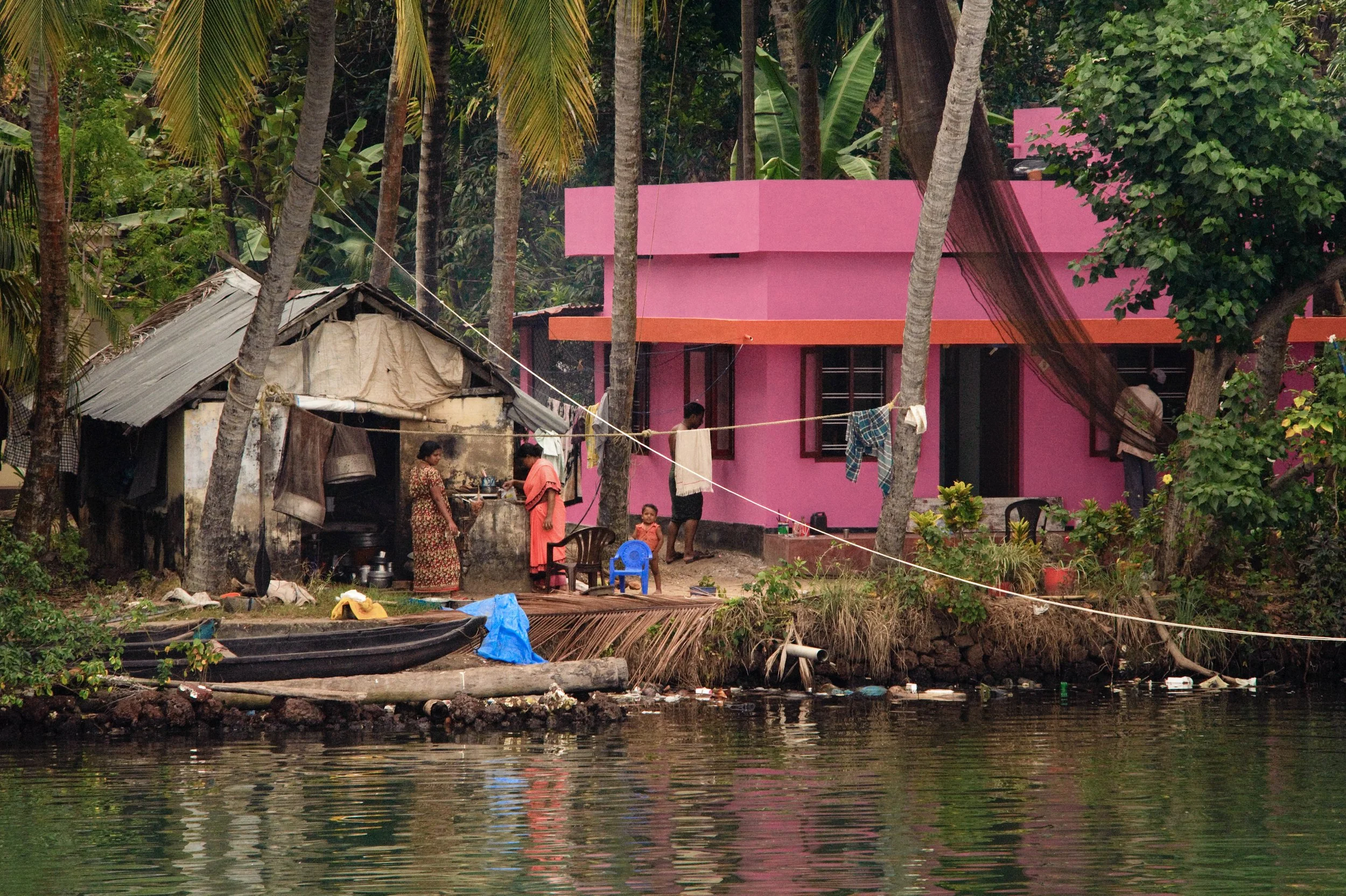 A group of people near a pink house with pink walls and black windows, surrounded by tall trees, next to a body of water with some trash, a small boat, and a makeshift shelter.