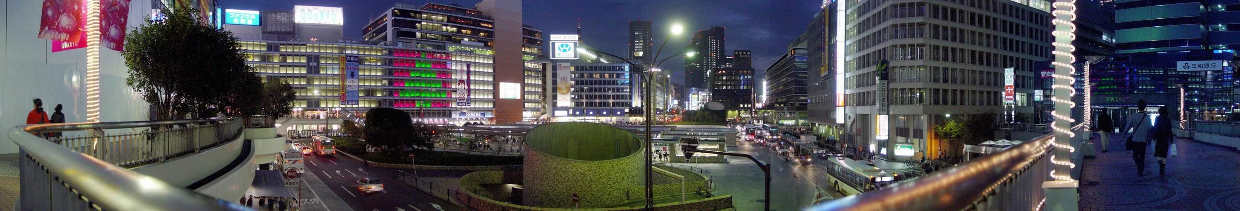 Nighttime cityscape view from a pedestrian bridge, featuring lit buildings, traffic on the streets, and people walking.