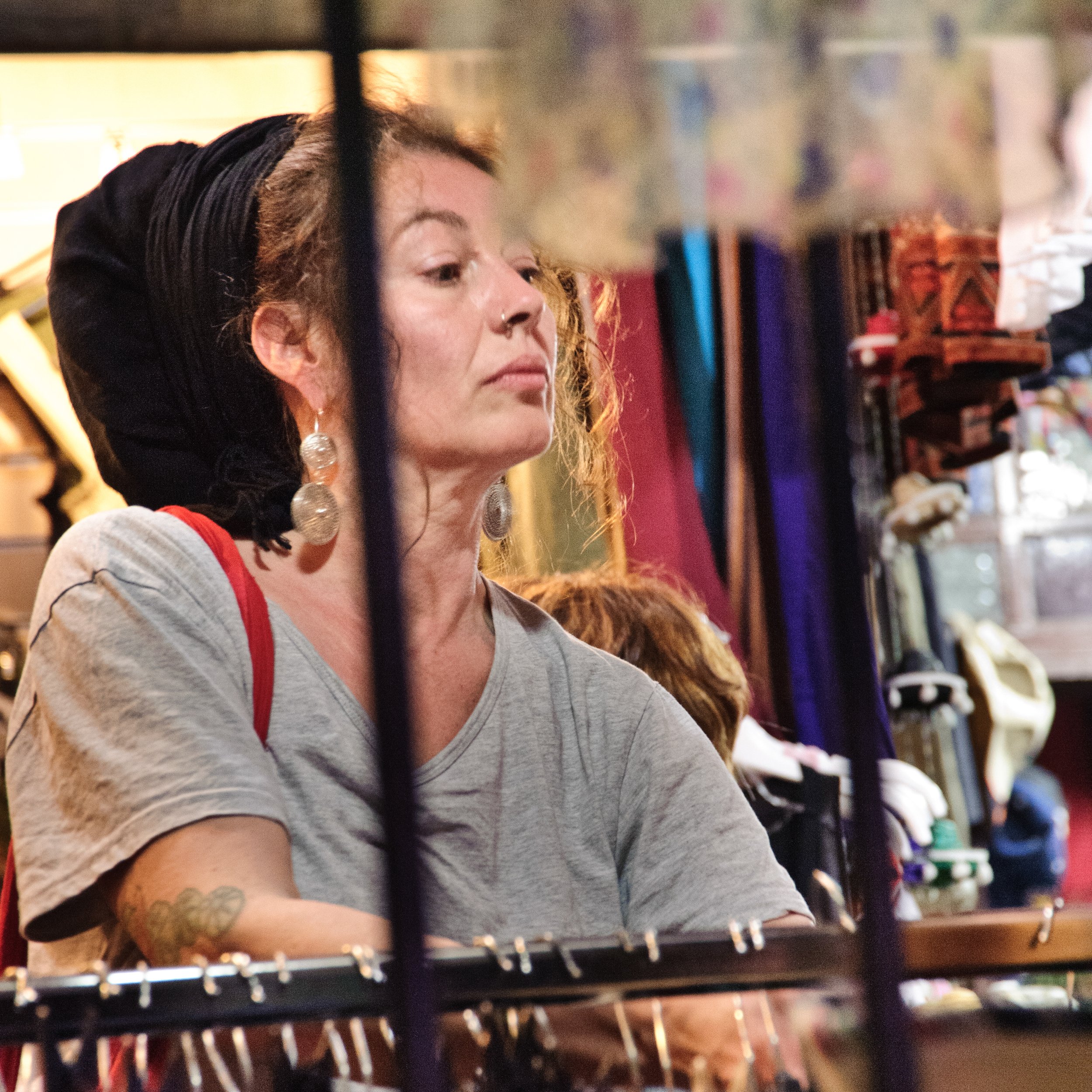 A woman with curly hair, large earrings, and a nose ring is browsing in a store, seen through a metal grid or rack. The background is filled with colorful items and shelves.