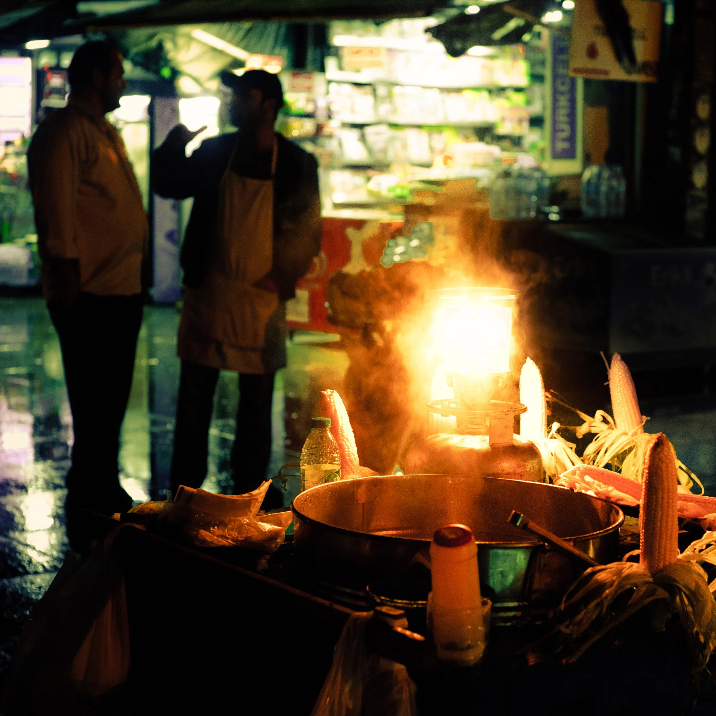 Street food stall at night with a small stove emitting flames, surrounded by corn cobs and cooking utensils, with two people conversing in the background.