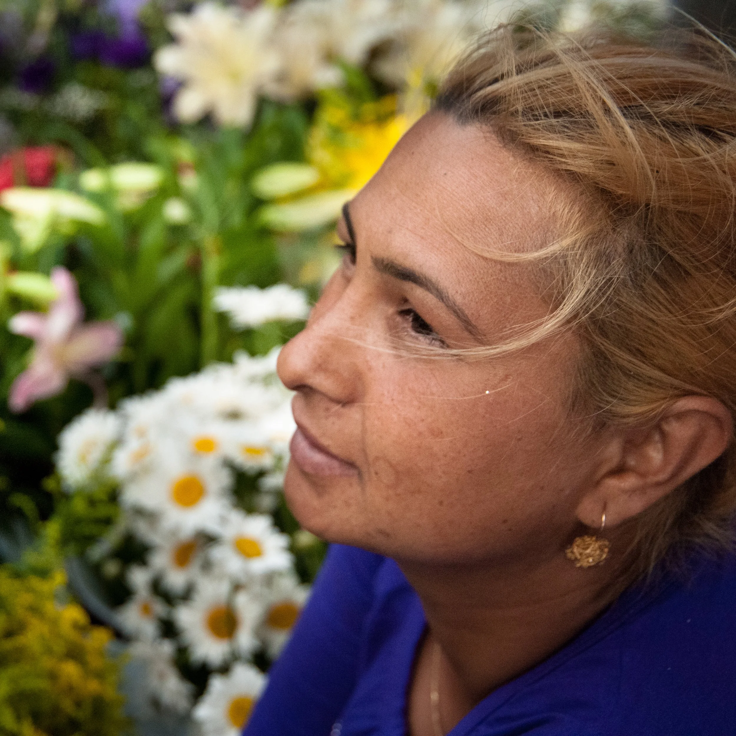 A woman with light skin and freckles, with blonde hair, wearing a blue top and gold earrings, surrounded by colorful flowers.