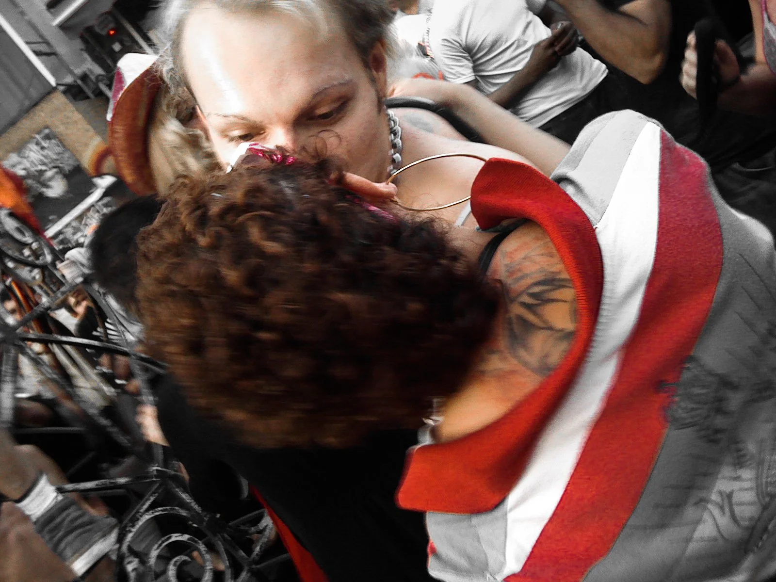 A woman with blonde hair kissing another person with curly red hair and a tattoo on their shoulder, in a crowded setting.