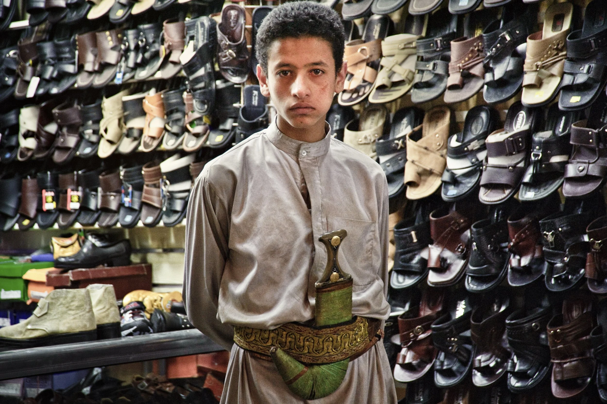 Young man in traditional Middle Eastern attire stands in front of a wall of sandals in a shoe store.
