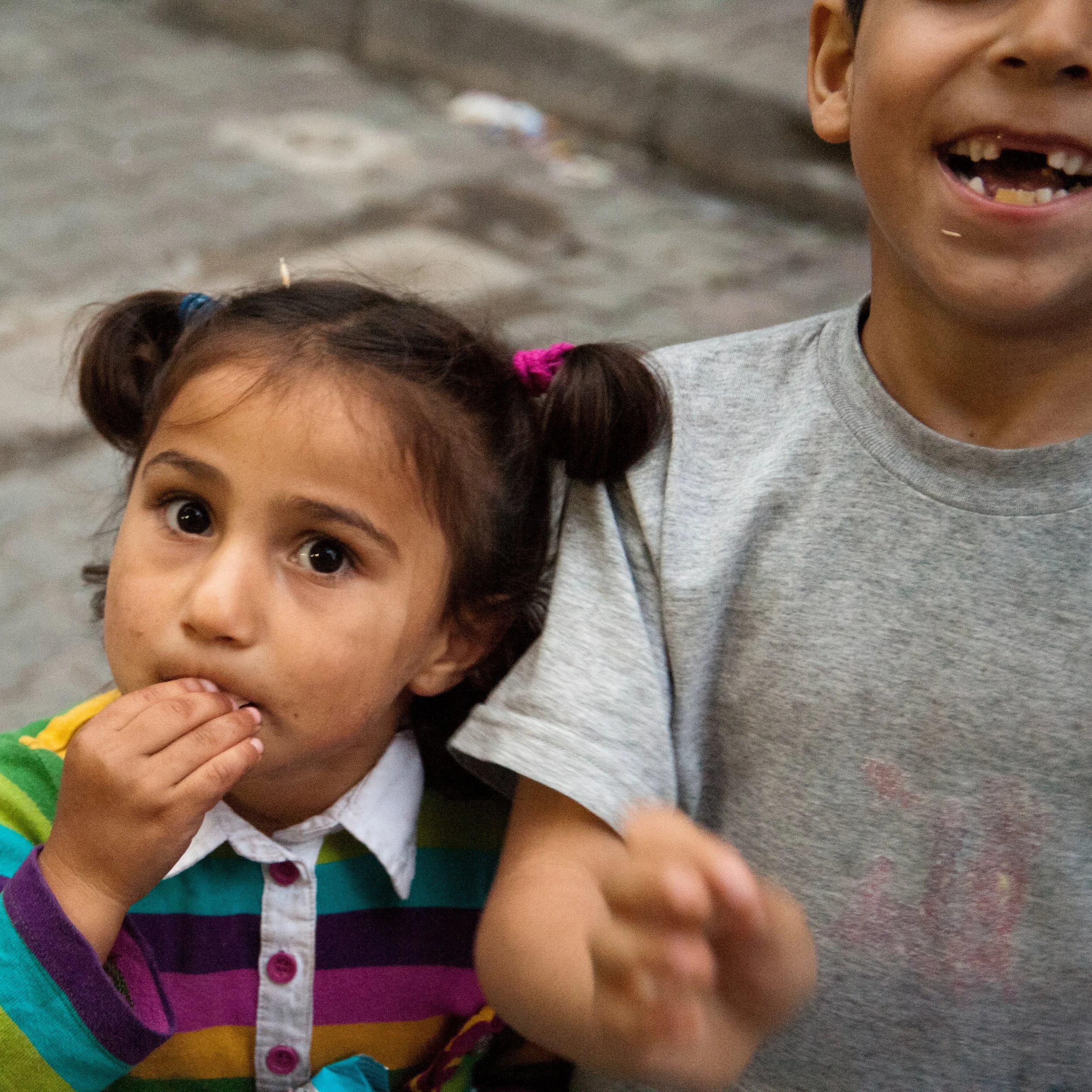 A young girl with dark hair in pigtails and big eyes, wearing a colorful striped shirt, has her hand near her mouth. Part of a smiling boy in a gray shirt with missing teeth is also visible, with the background showing a sandy outdoor setting.