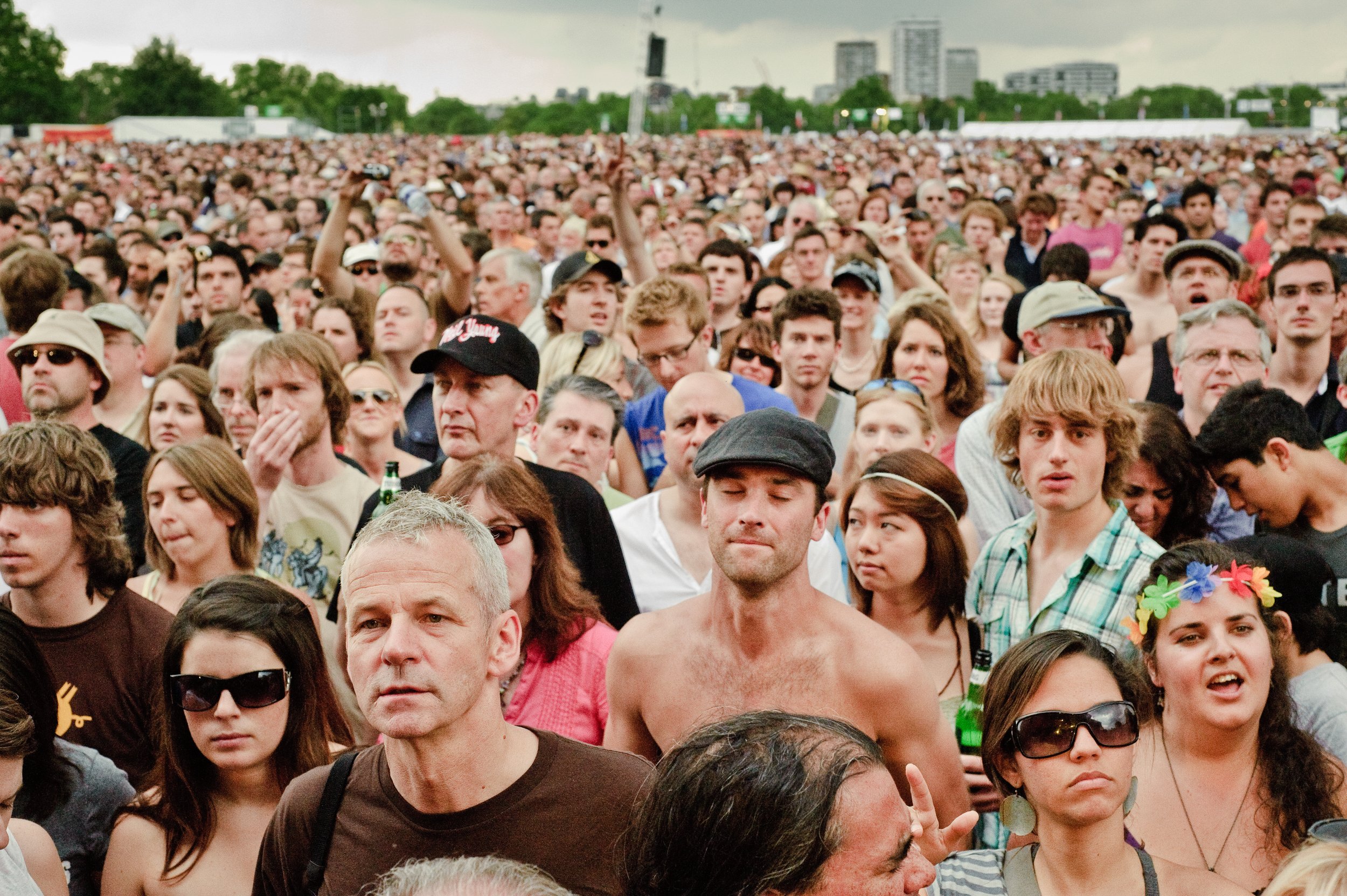 A large crowd of people at an outdoor concert or festival with trees and city buildings in the background.
