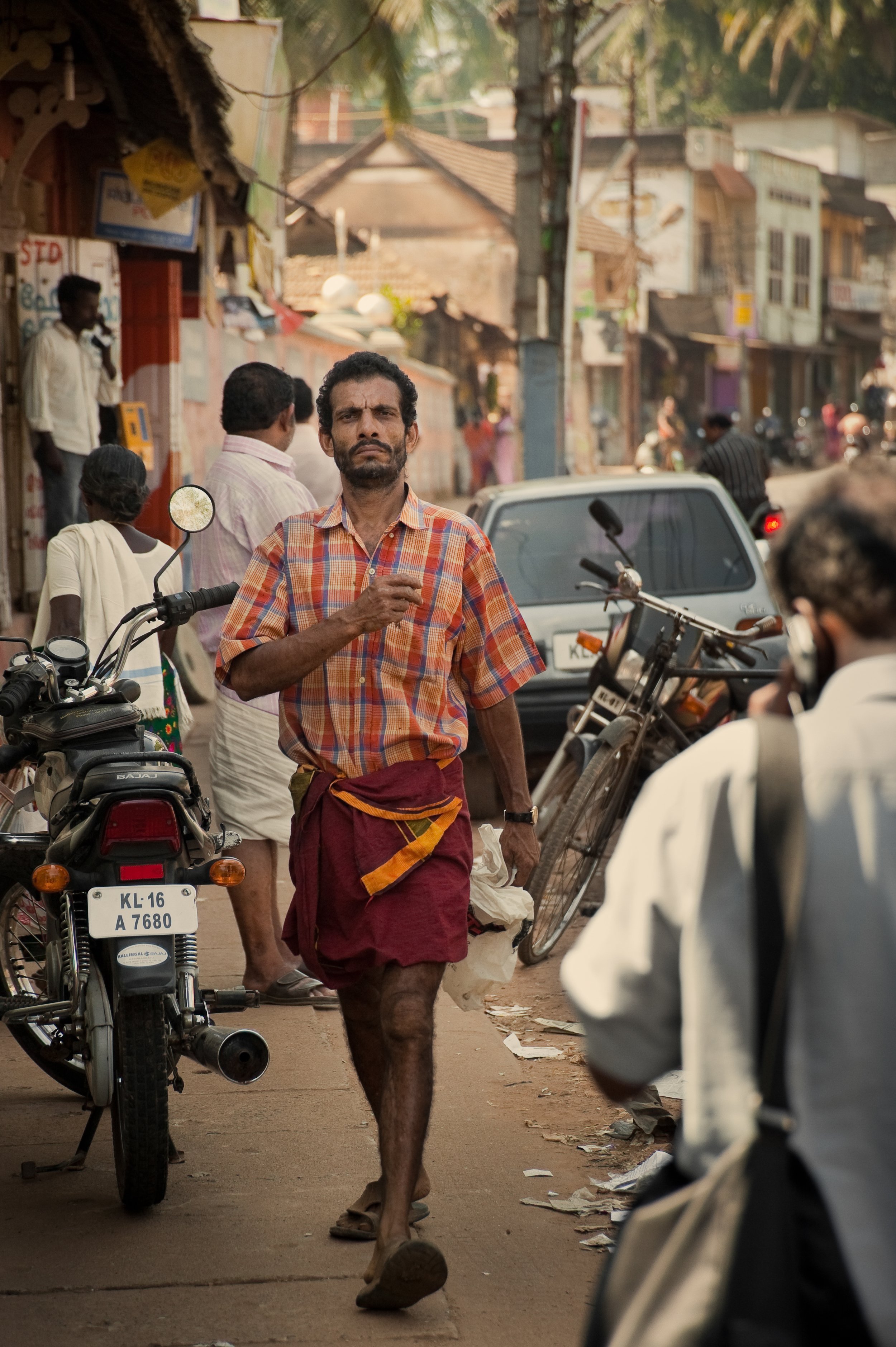 A man wearing a checkered shirt and a traditional Indian dhoti walks down a busy street, holding a white bag in one hand and gesturing with the other. The street is crowded with people, motorcycles, bicycles, and cars, with small shops and buildings 