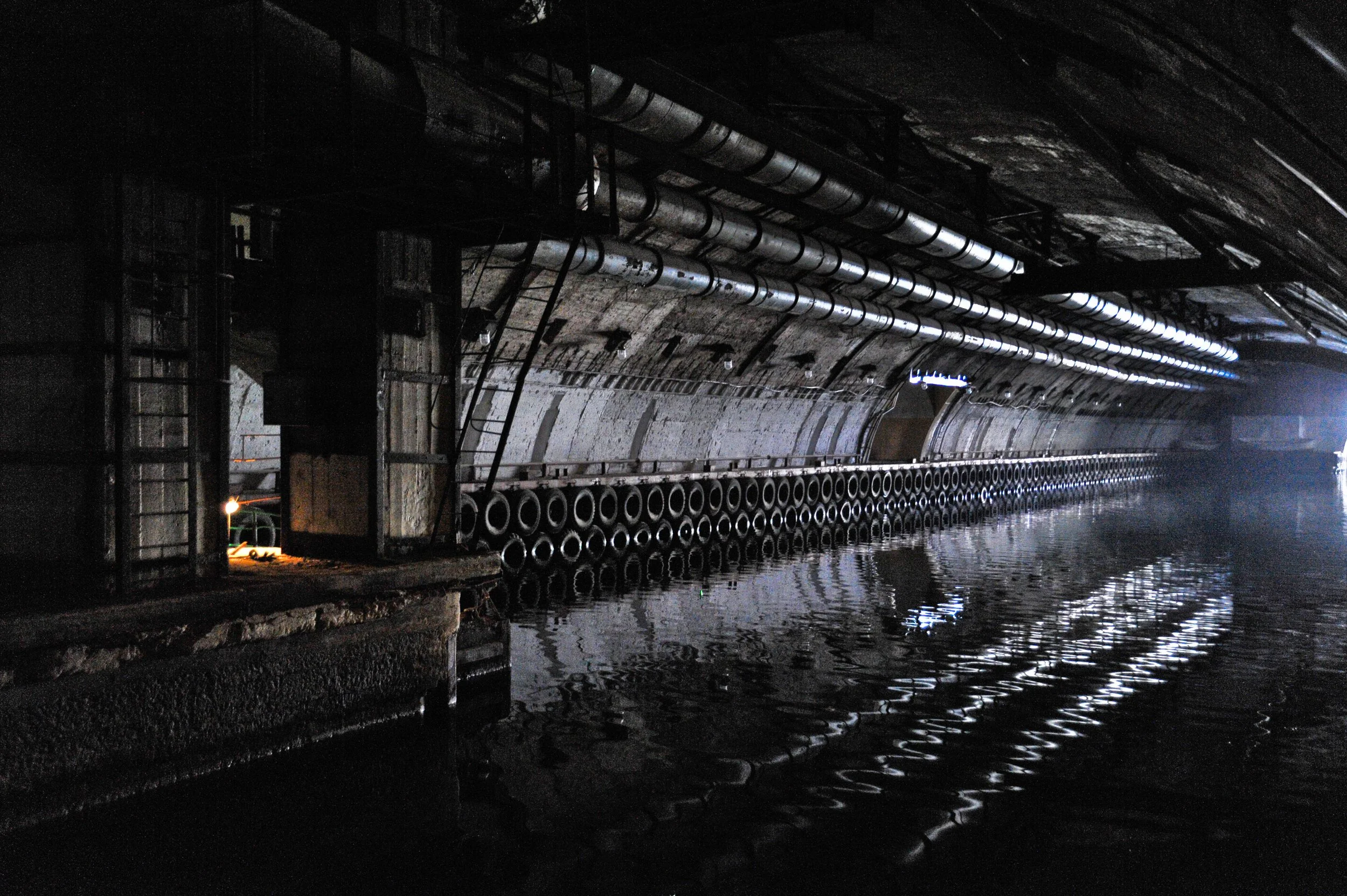 Underground tunnel with sewer pipes and water at the bottom, dimly lit with reflections on water surface.