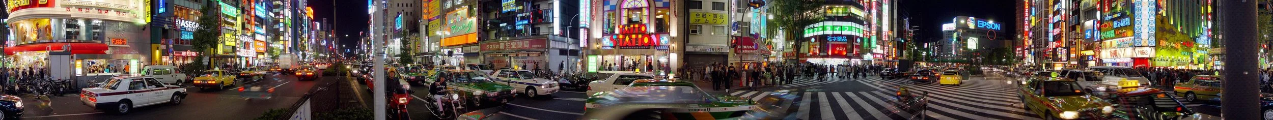 Nighttime busy city street filled with cars, taxis, motorcycles, and pedestrians, with bright neon and electronic billboards on buildings.
