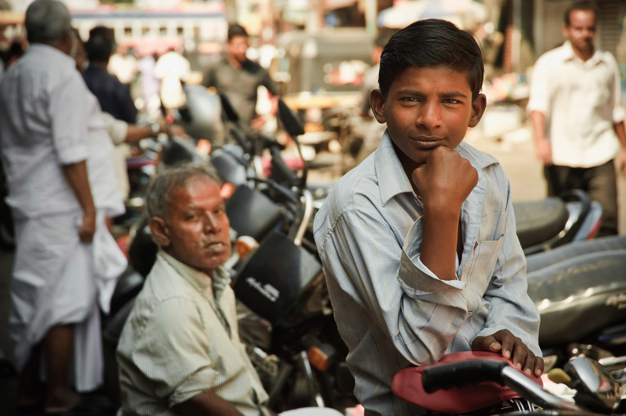 Young boy in a light-colored shirt sitting on a red motorcycle, with an older man and other people in the background on a busy street.