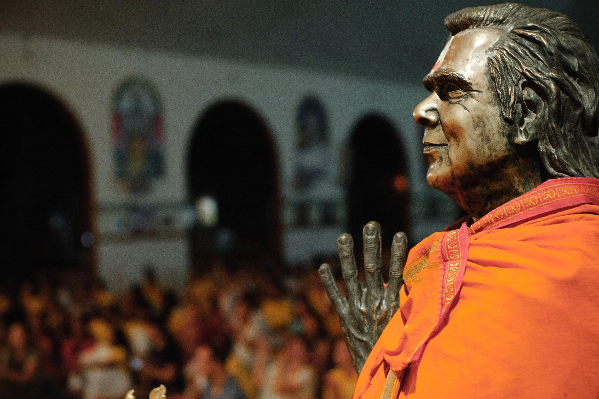 Close-up of a bronze statue of a man with flowing hair, wearing an orange shawl, with hands clasped in a prayer or greeting gesture, in a church or shrine filled with people.