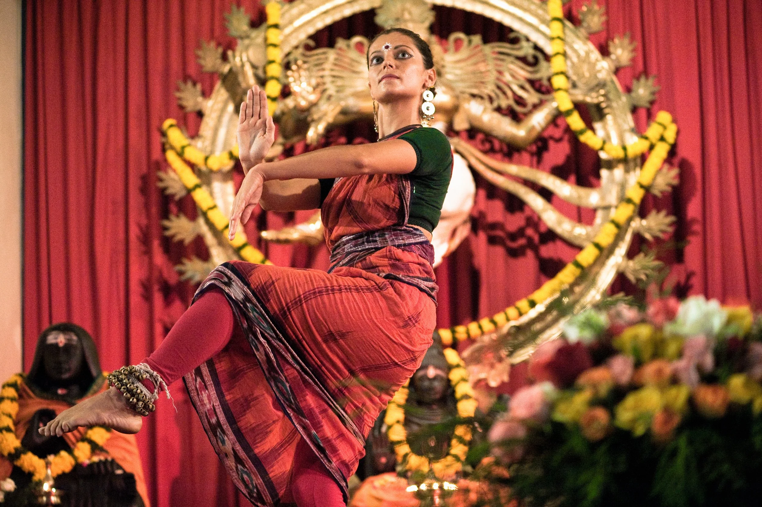 A woman performing a traditional Indian dance on stage, with a decorative backdrop featuring a large idol and yellow flower garlands, dressed in a red and black saree with jewelry.
