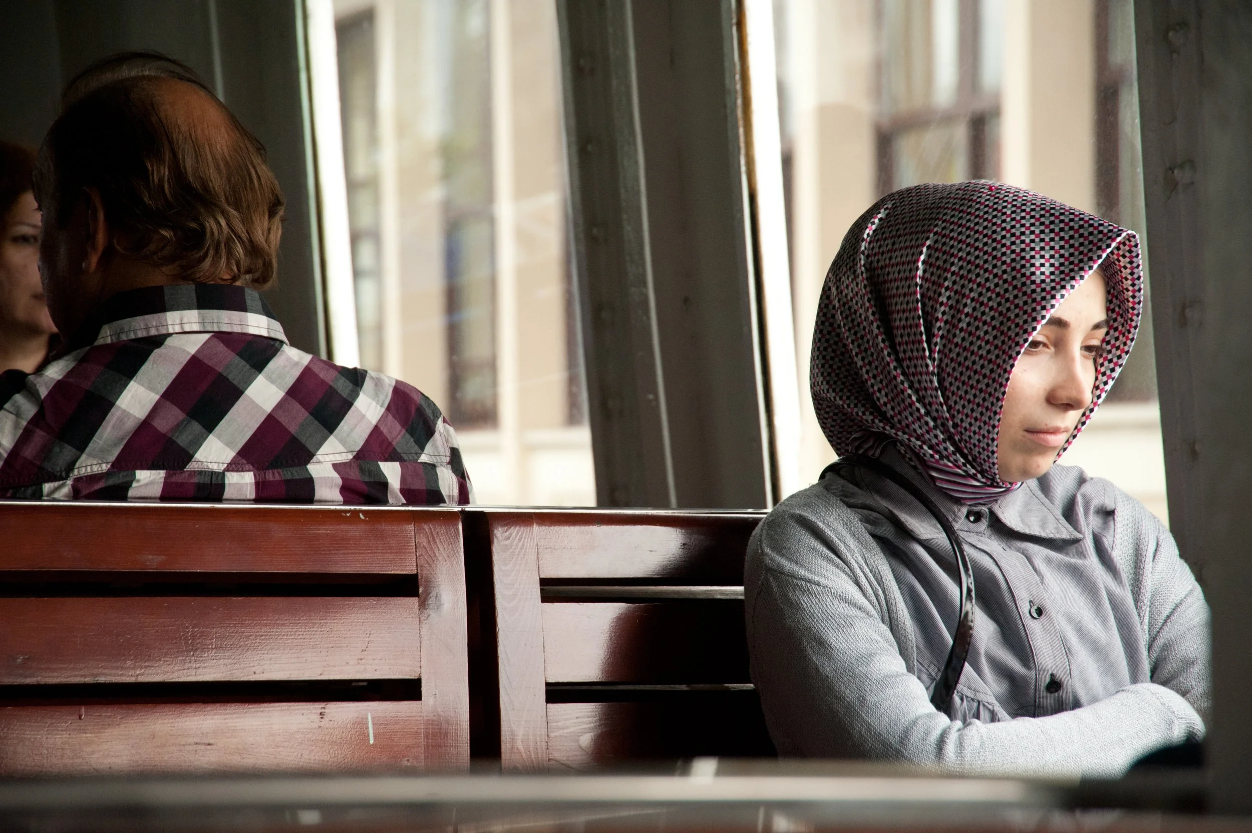 A young woman wearing a checkered headscarf sitting alone on a wooden bench inside a cafe, looking contemplative with eyes closed or downcast, while a man with short hair and a checkered shirt sits nearby with his back to the camera.