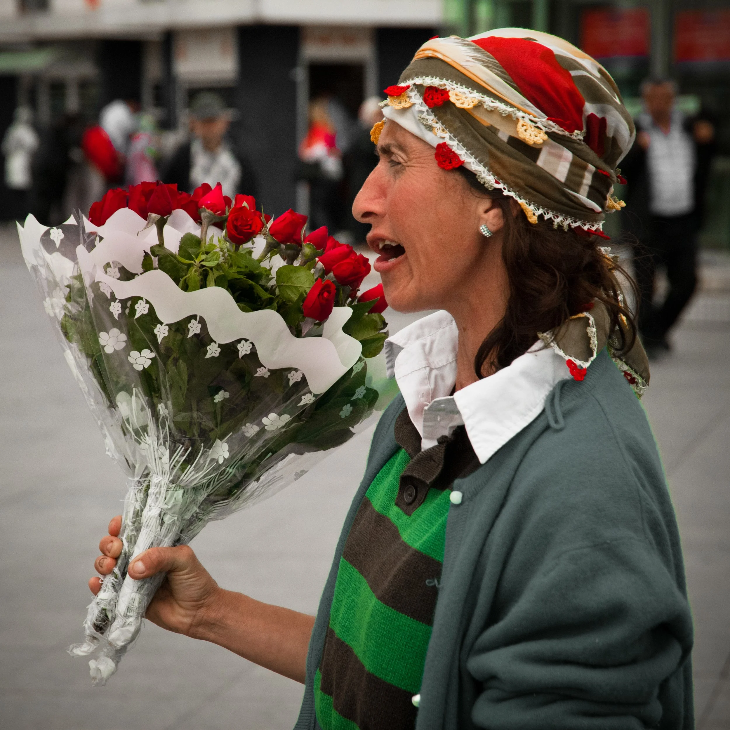 A woman with a patterned headscarf holding a bouquet of red roses, speaking or singing, in an outdoor public setting.