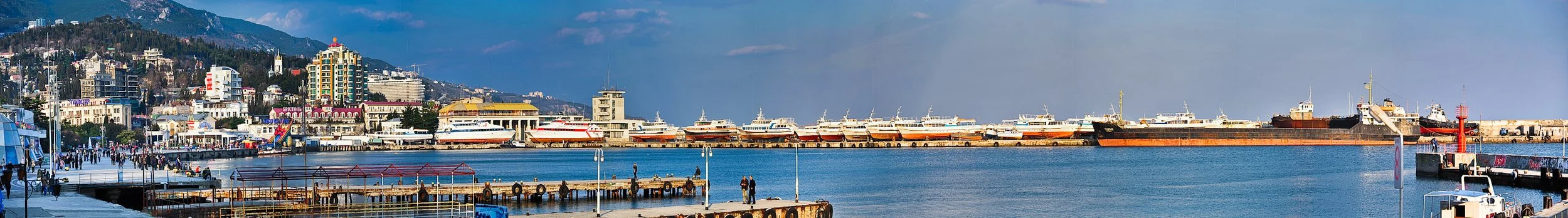 Panoramic view of a harbor with multiple boats docked, a pier with people, and a city skyline with colorful buildings and mountains in the background.