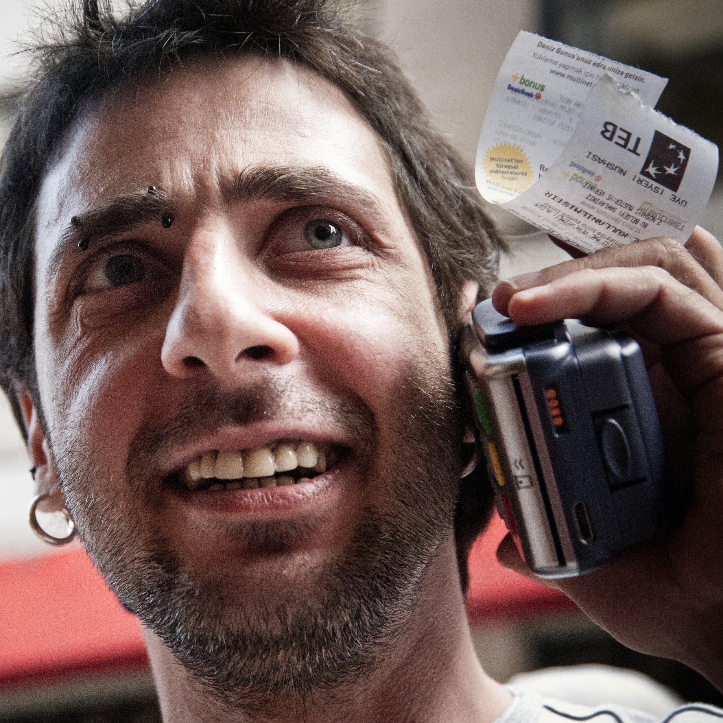Close-up of a smiling man with braces on his teeth, a piercing above his right eyebrow, earrings, and facial hair, talking on a cell phone while holding a receipt in his hand.