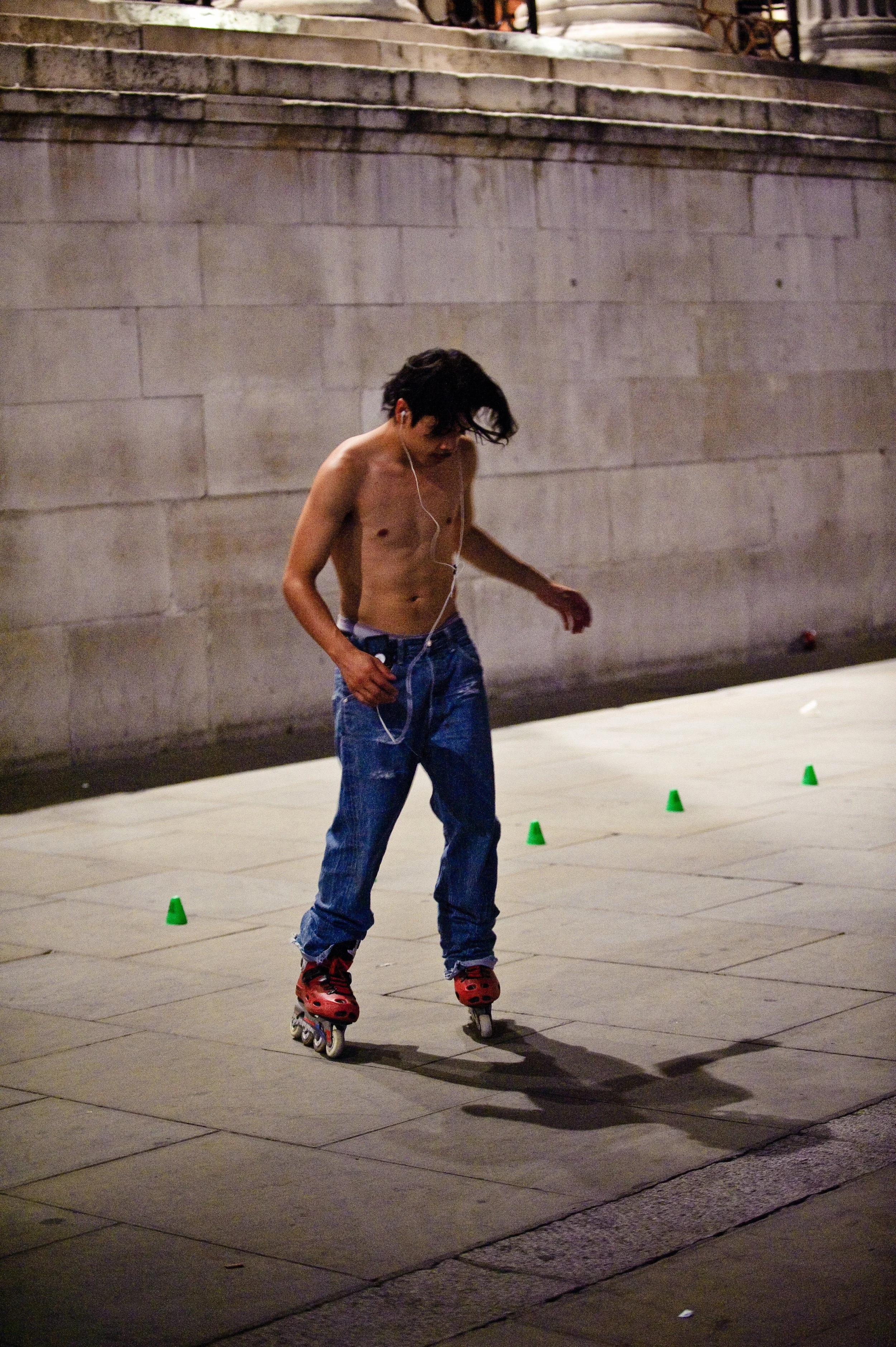 A shirtless young man with long dark hair skating on a city sidewalk at night, with four small green cones arranged behind him.