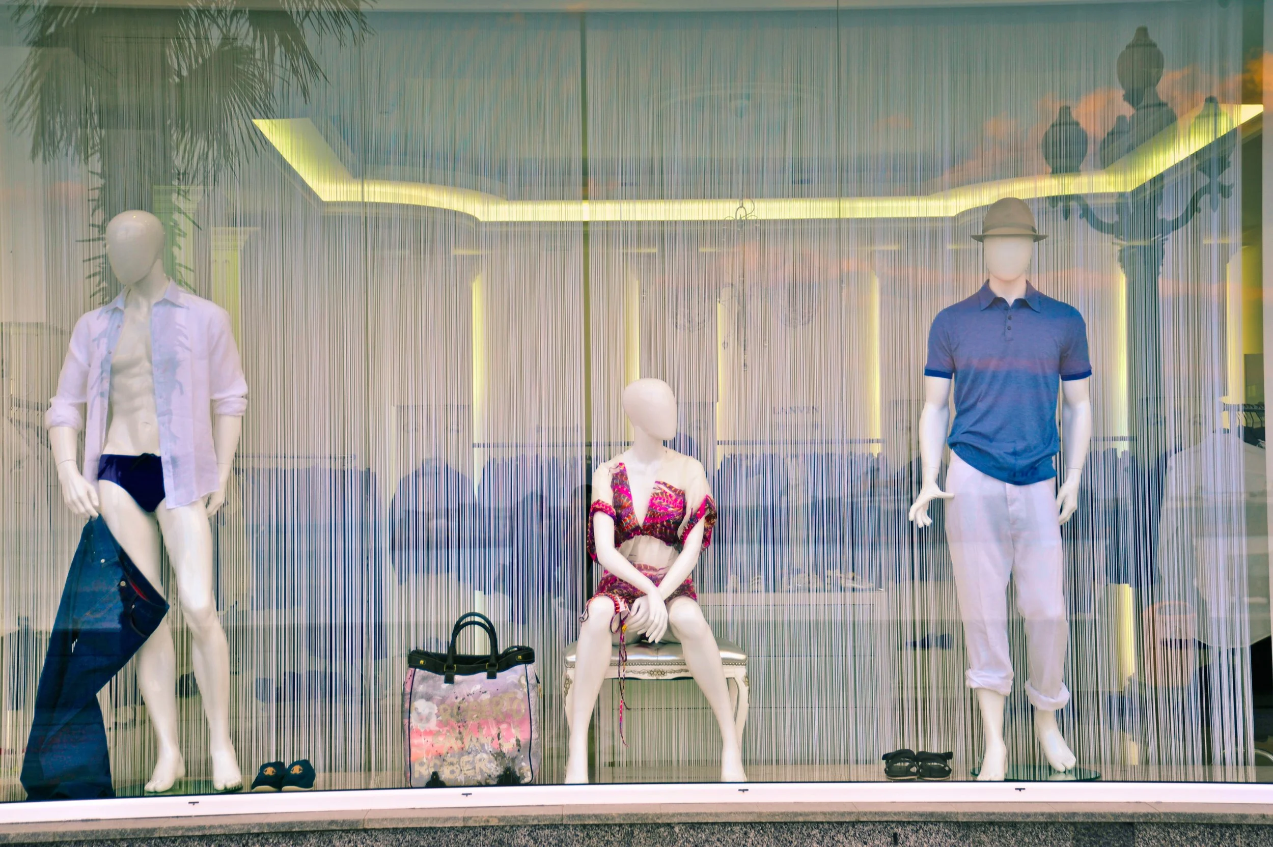 Store window display with mannequins dressed in summer clothing, one sitting and two standing, with accessories and bags placed in front.