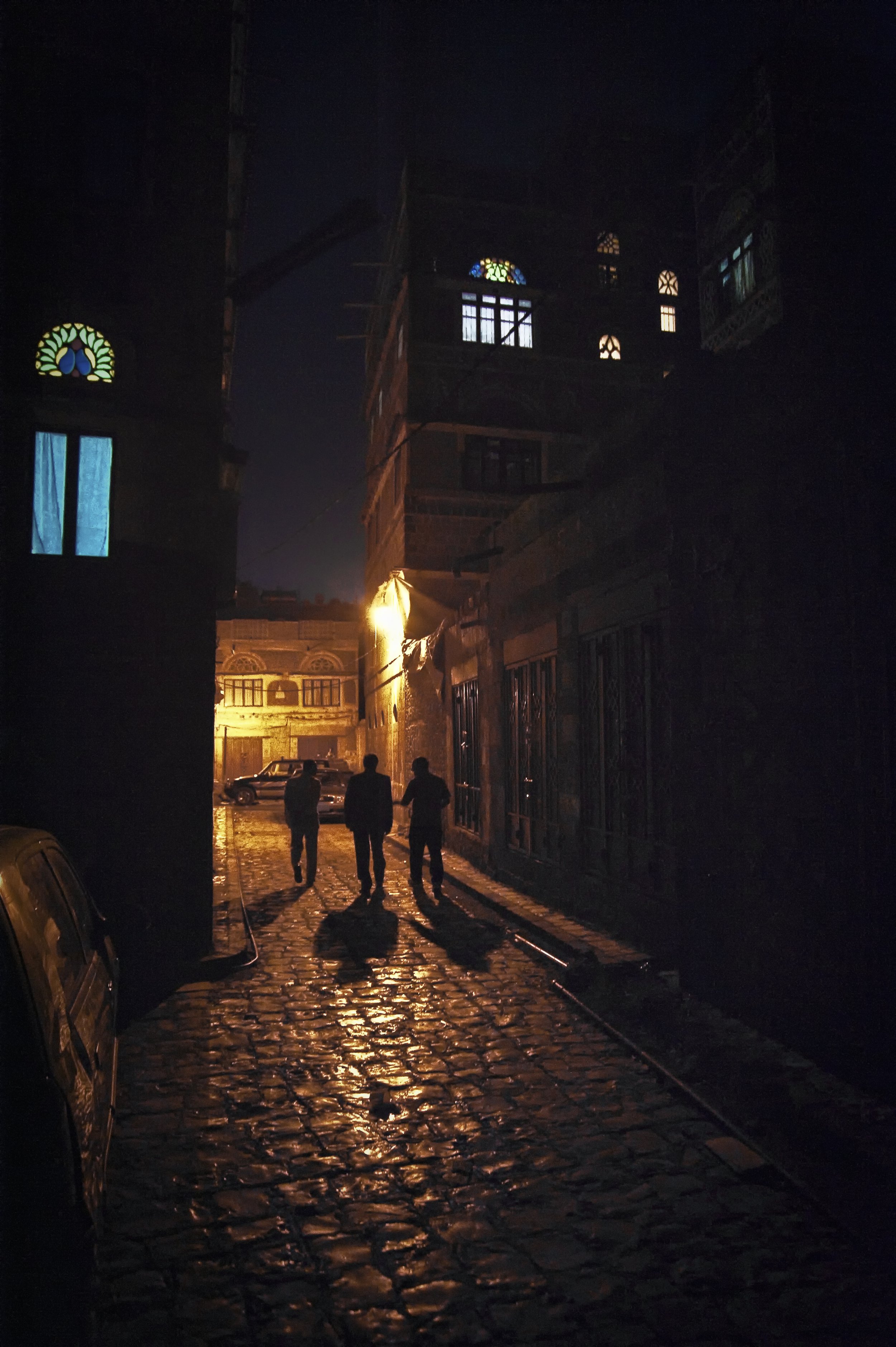 Three people walking down a cobblestone street at night with shadows cast by streetlights, buildings with stained glass windows, and parked cars.
