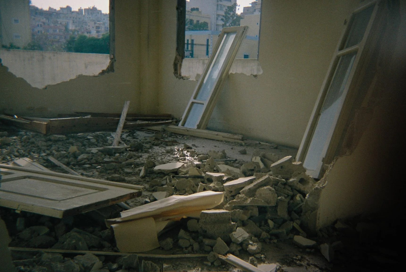 Room with large broken holes in the walls, debris on the floor, and leaning window frames, suggesting recent destruction or construction.