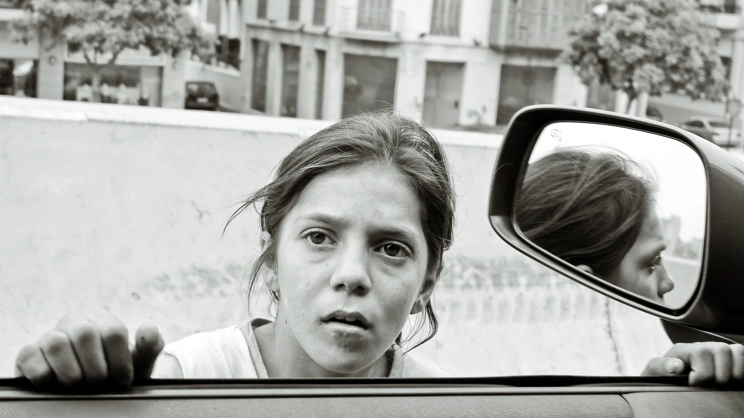 A young girl looking through the open window of a car, with her hand gripping the window edge, and her reflection visible in the side mirror.
