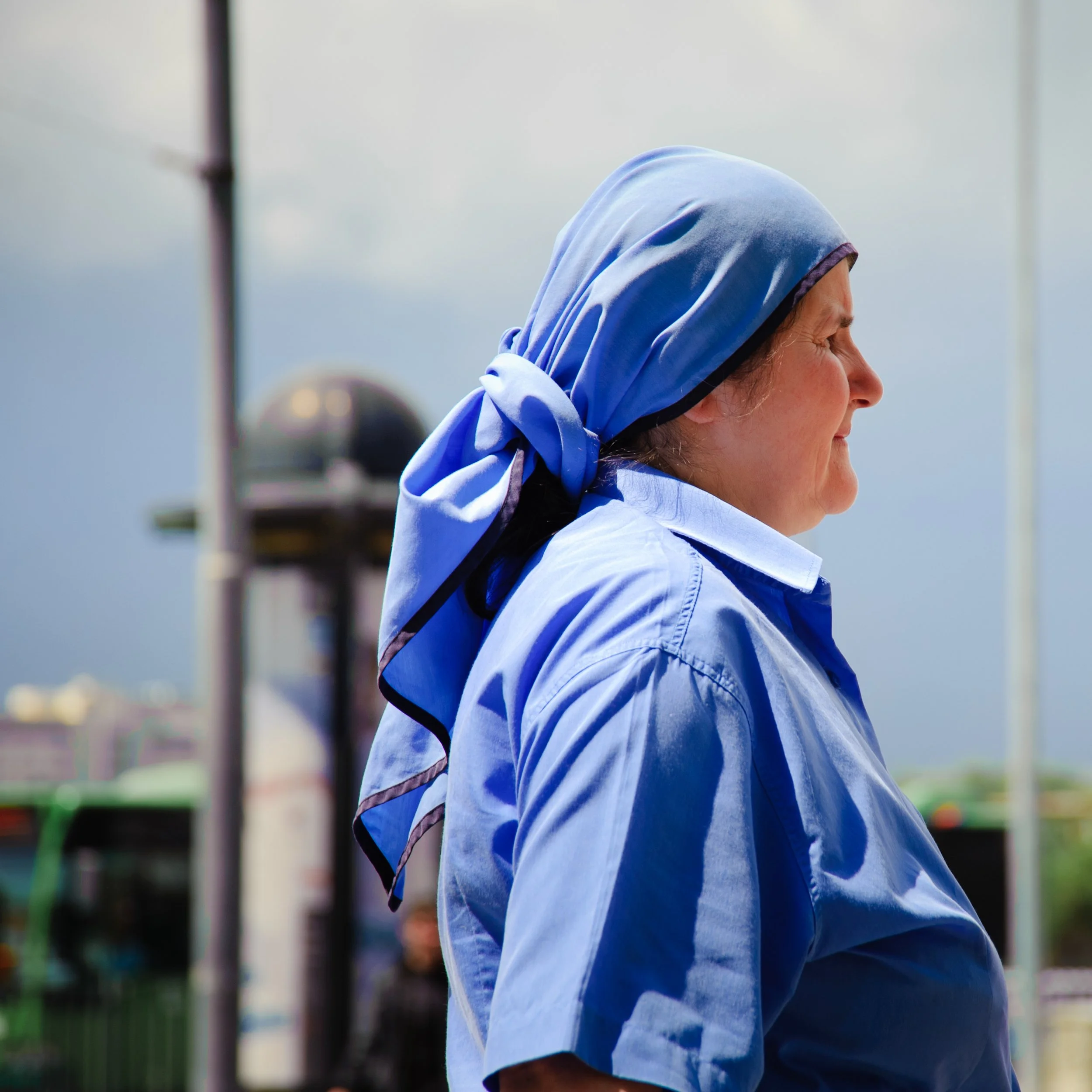 A woman in profile wearing a blue headscarf and blue shirt outdoors with a cloudy sky and buses in the background.