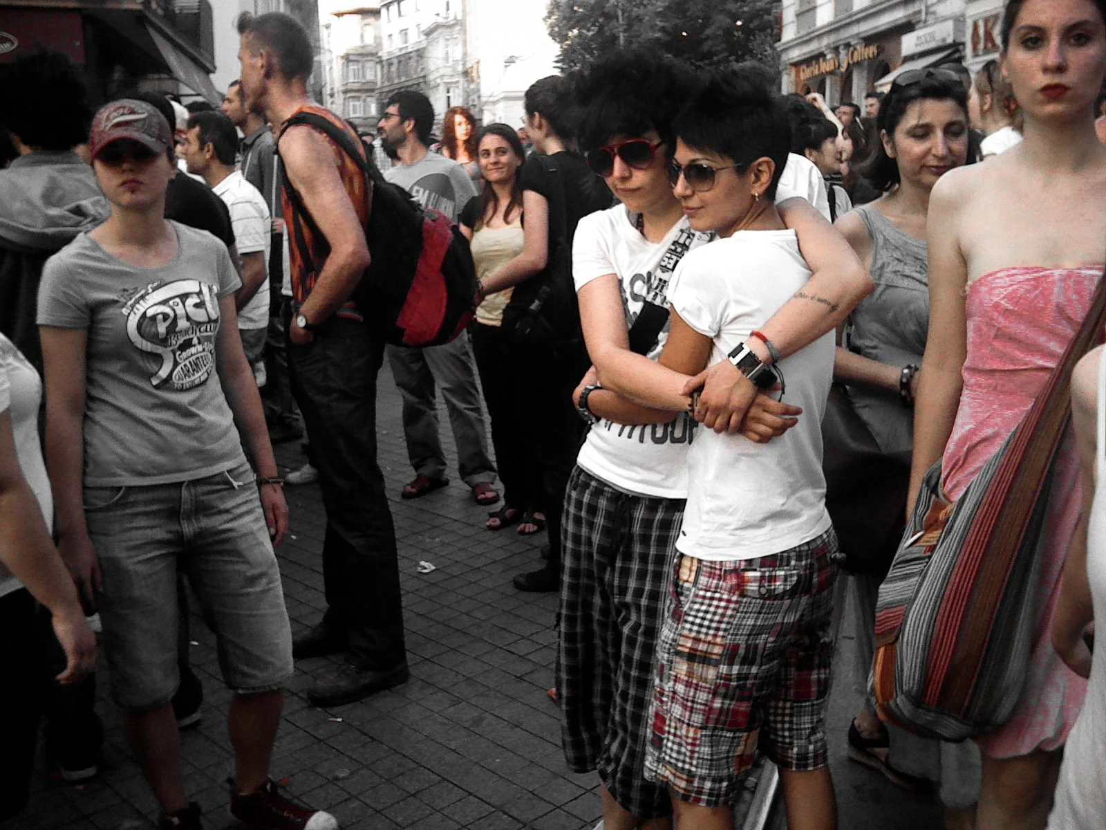 A crowd of people standing in a city street. Two women are hugging in the center, one wearing sunglasses and the other with dark, short hair. Others are standing nearby, some looking at the camera, while some are looking elsewhere. The background sho