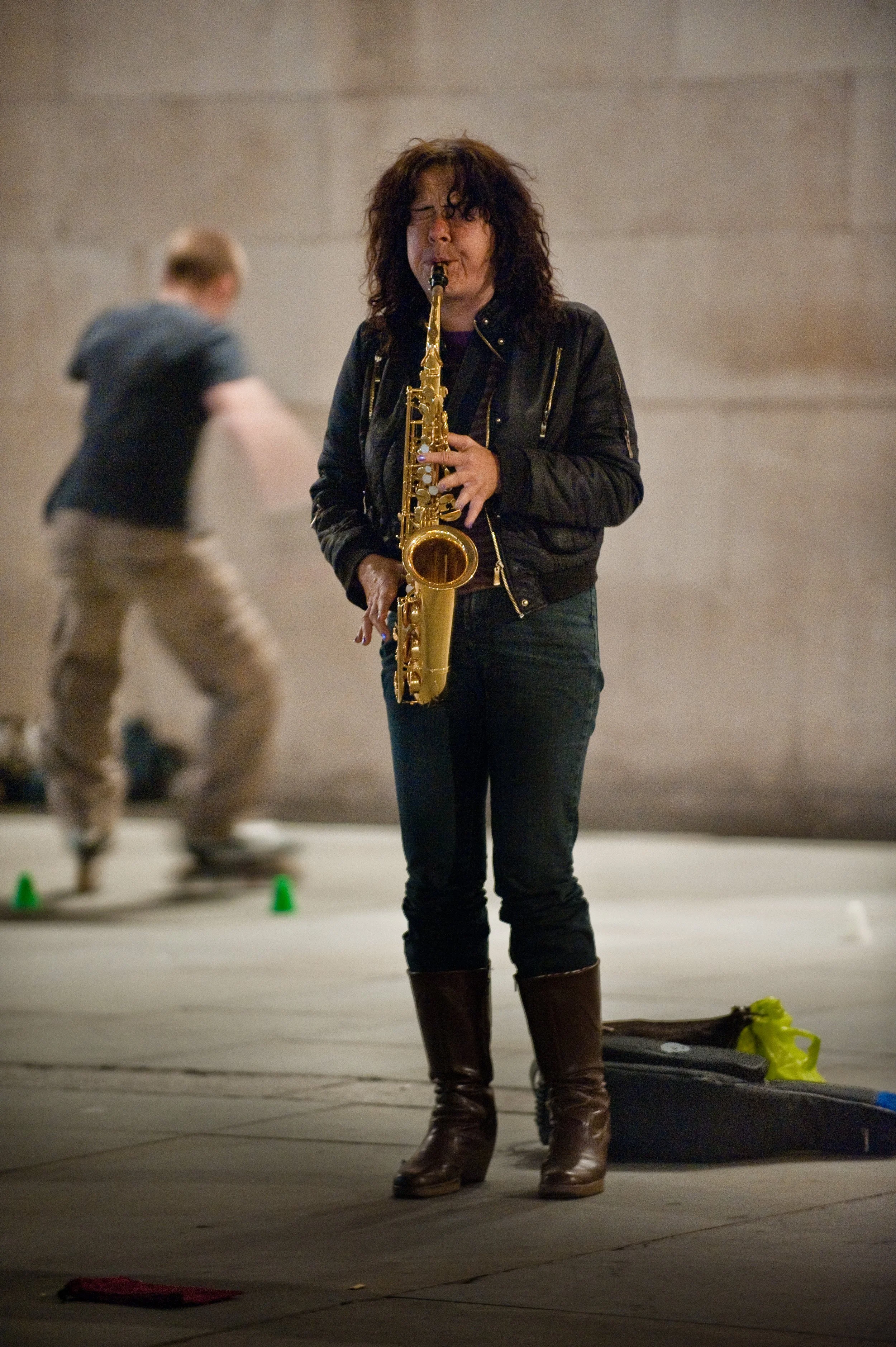Woman playing a saxophone on the street while a man skateboards in the background.