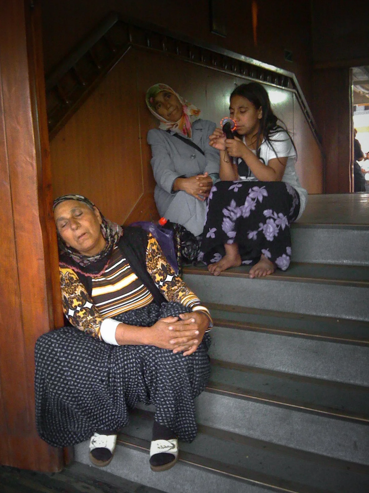 A woman rests on the stairs with her eyes closed, wearing a striped top, patterned pants, and sandals. Two young women are sitting on the steps above her, one wearing a headscarf and the other playing a small musical instrument.