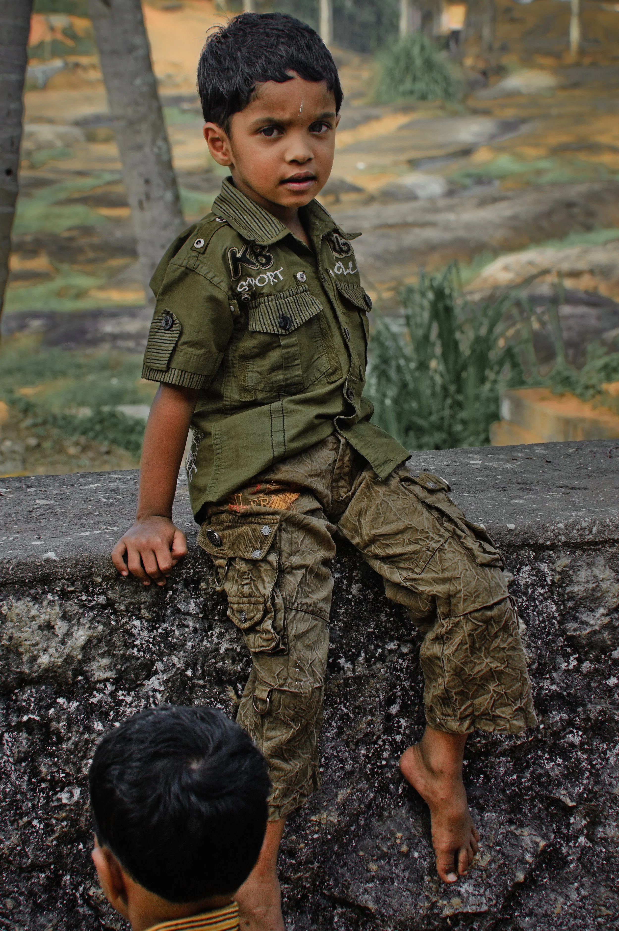 A young boy in military-style camouflage clothing sitting on a stone ledge outdoors.