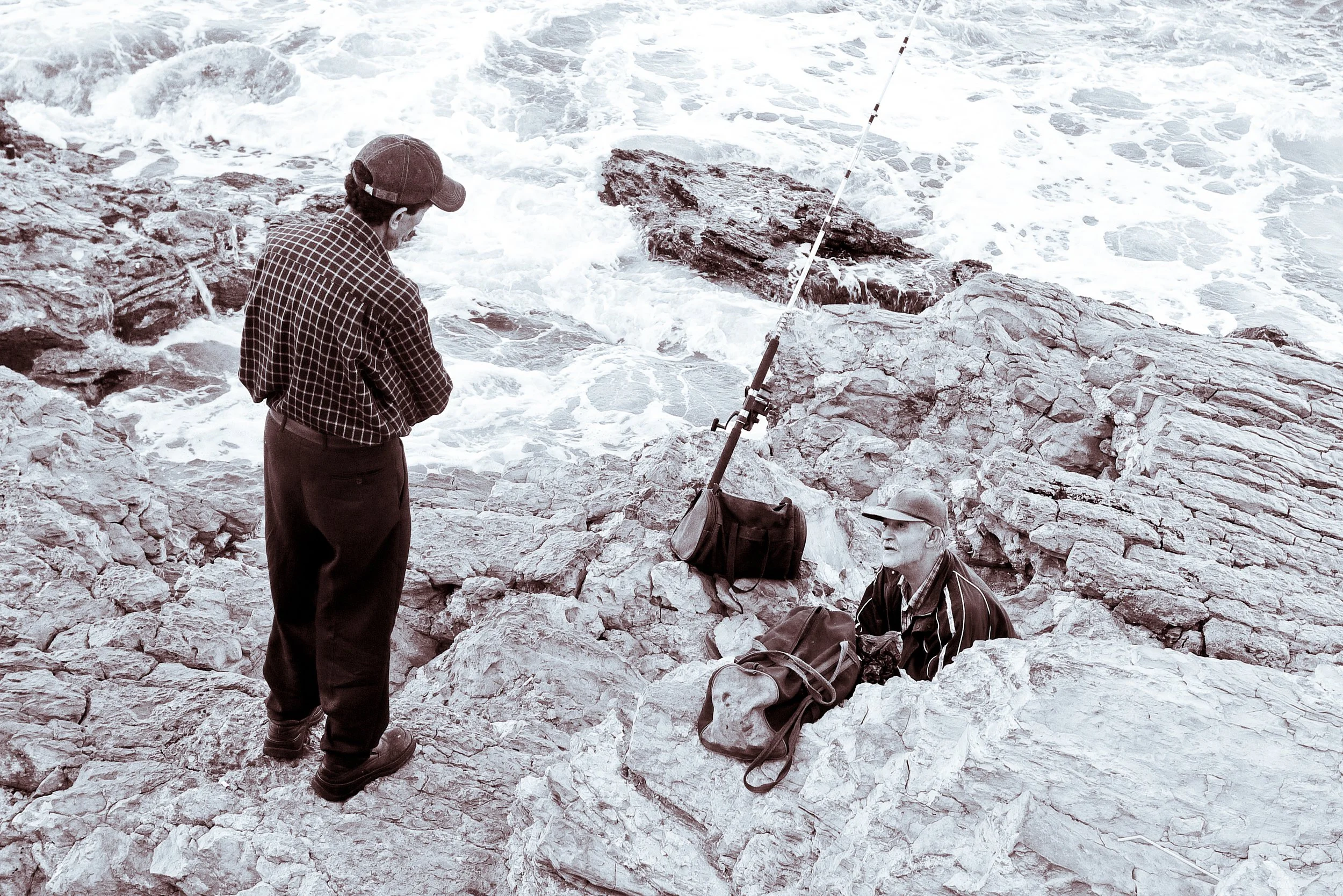 Two men on rocky shore, one standing and one sitting, near the ocean with fishing gear.