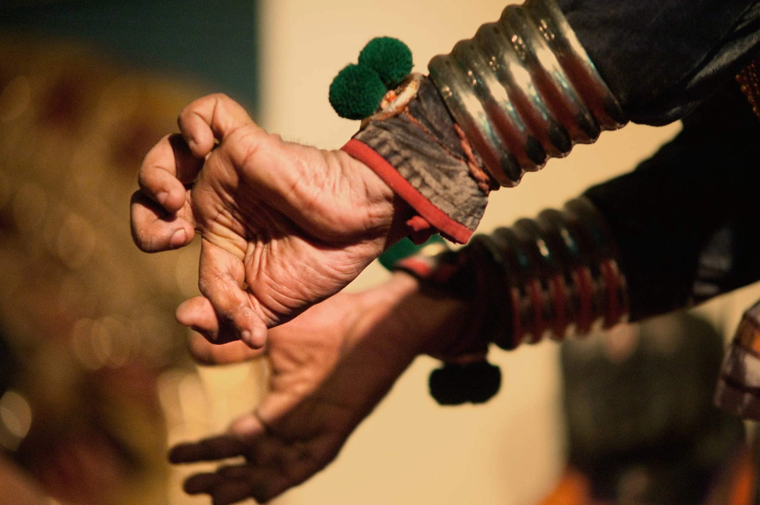 Close-up of two hands clasped together, one appearing elderly and worn, wearing colorful bracelets and a sweater sleeve, the other partially visible with a black sleeve.