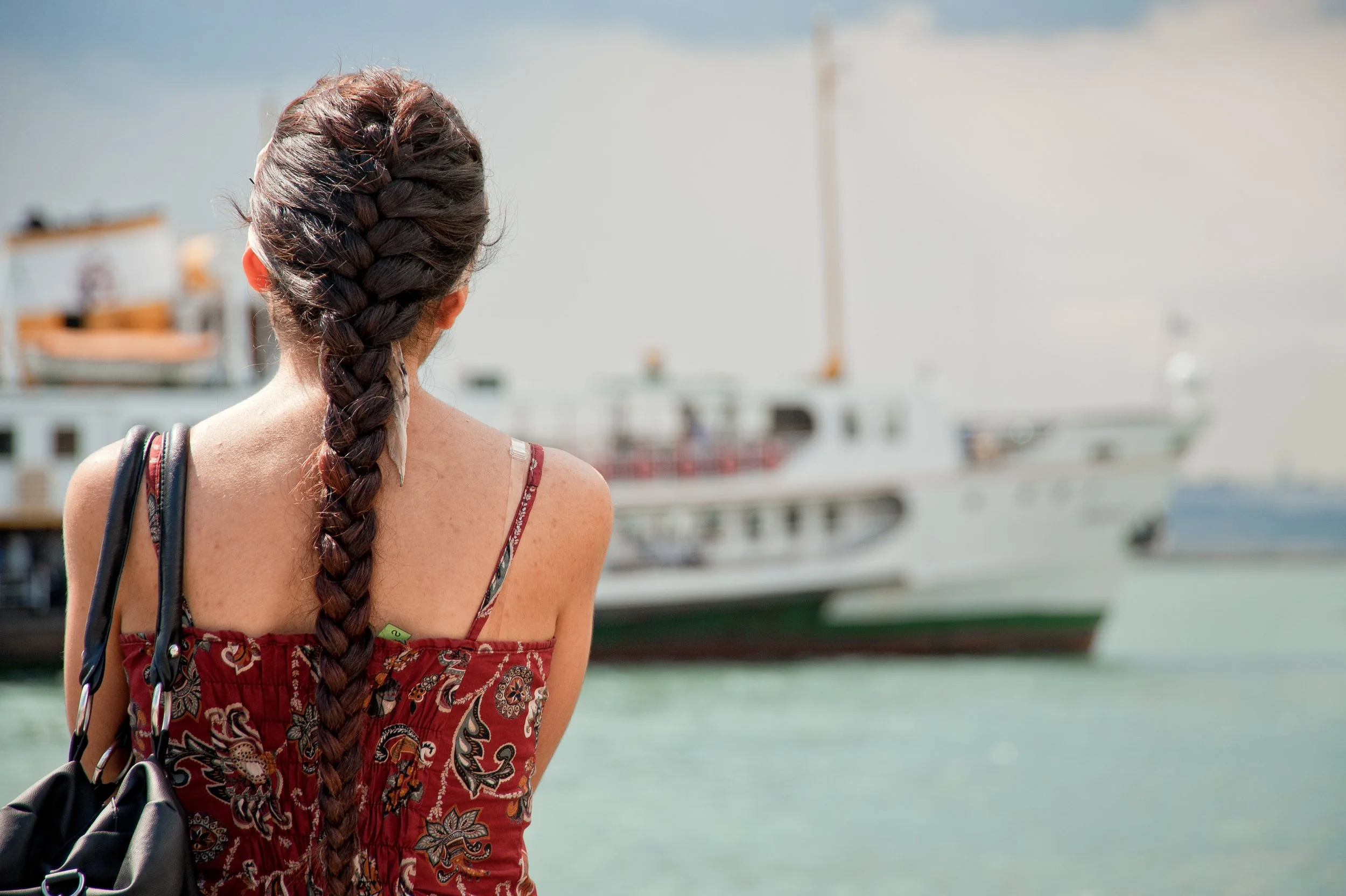 A woman with braided brown hair, wearing a red patterned dress, carrying a black bag, standing by the water with boats in the background.