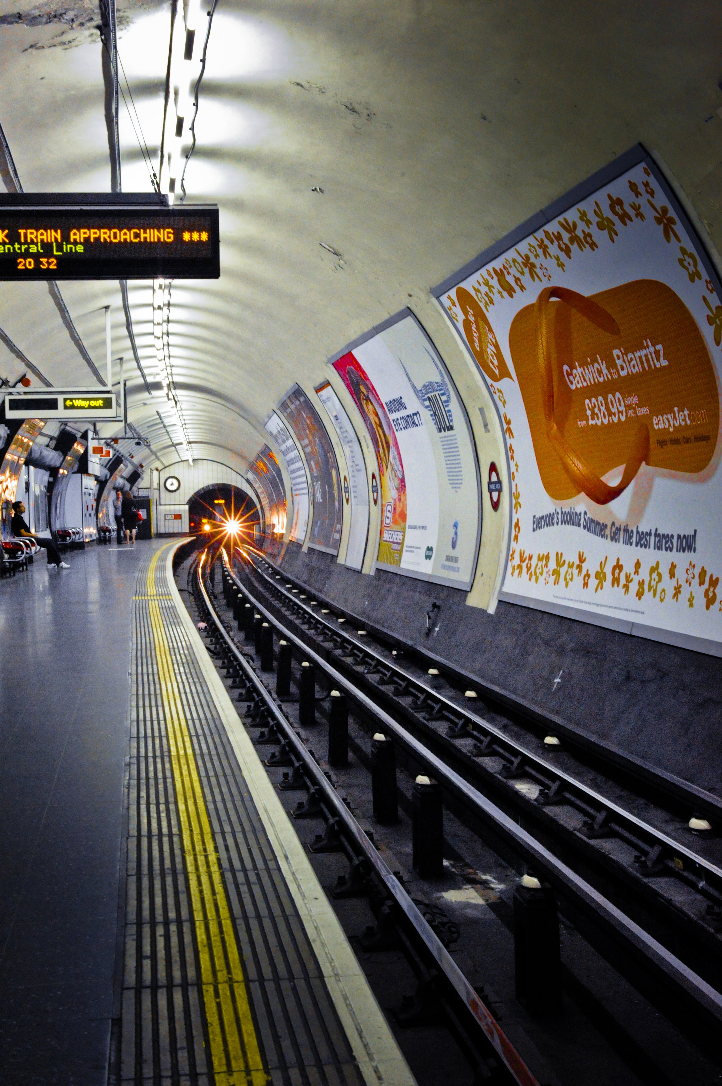 Subway station platform with train approaching, digital sign indicating train approaching, advertisements on the wall, and a few standing passengers.