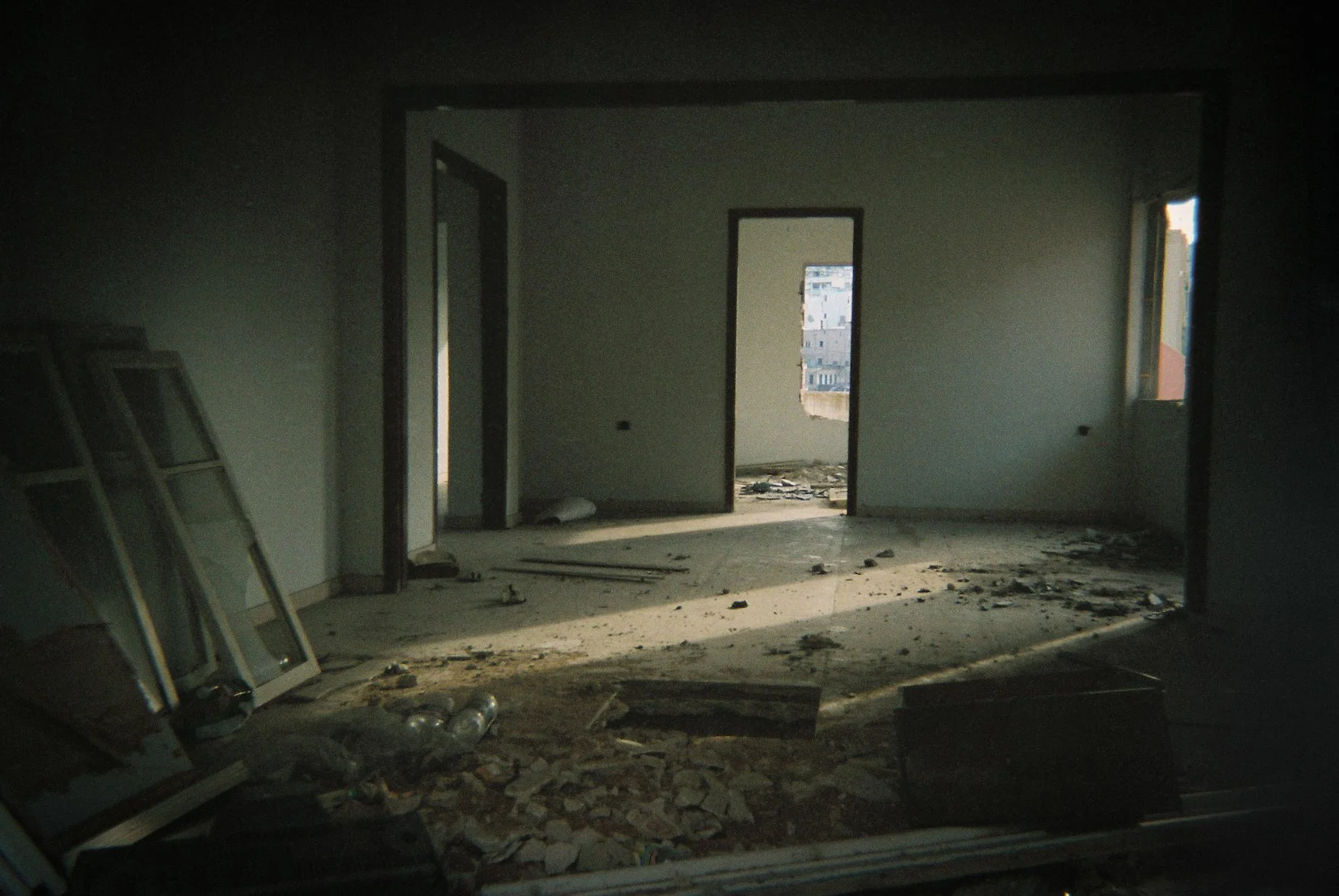 Interior of an unfinished or damaged building with debris on the floor, open doorways, and a window showing outside buildings.