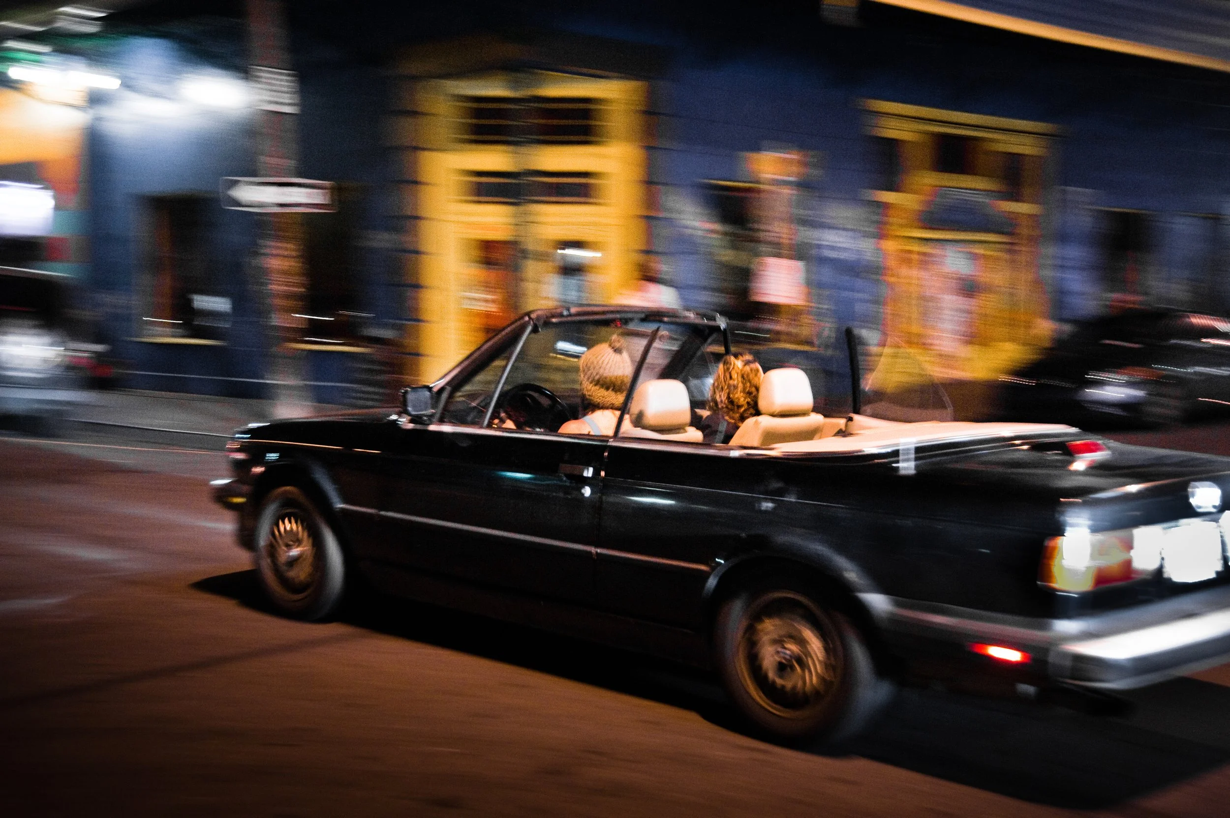 A black convertible car with beige interior driving through a city street at night, with colorful buildings and blurred lights in the background.