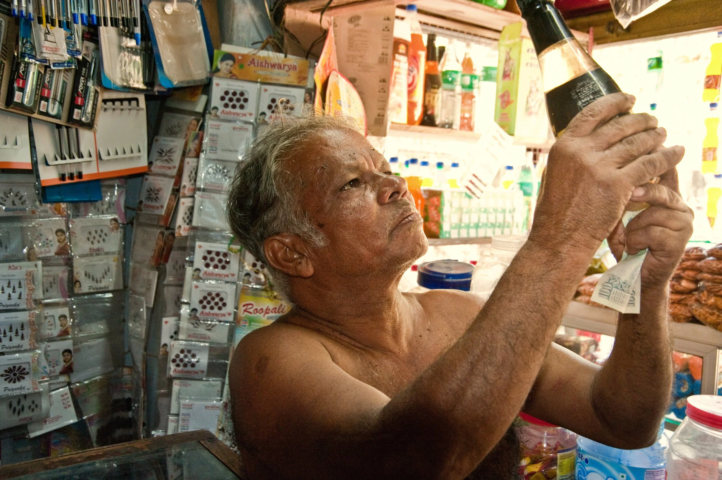 An elderly man with gray hair and a serious expression, shirtless, holding a bottle of dark-colored beverage inside a small shop with various household items and packaging on the shelves.