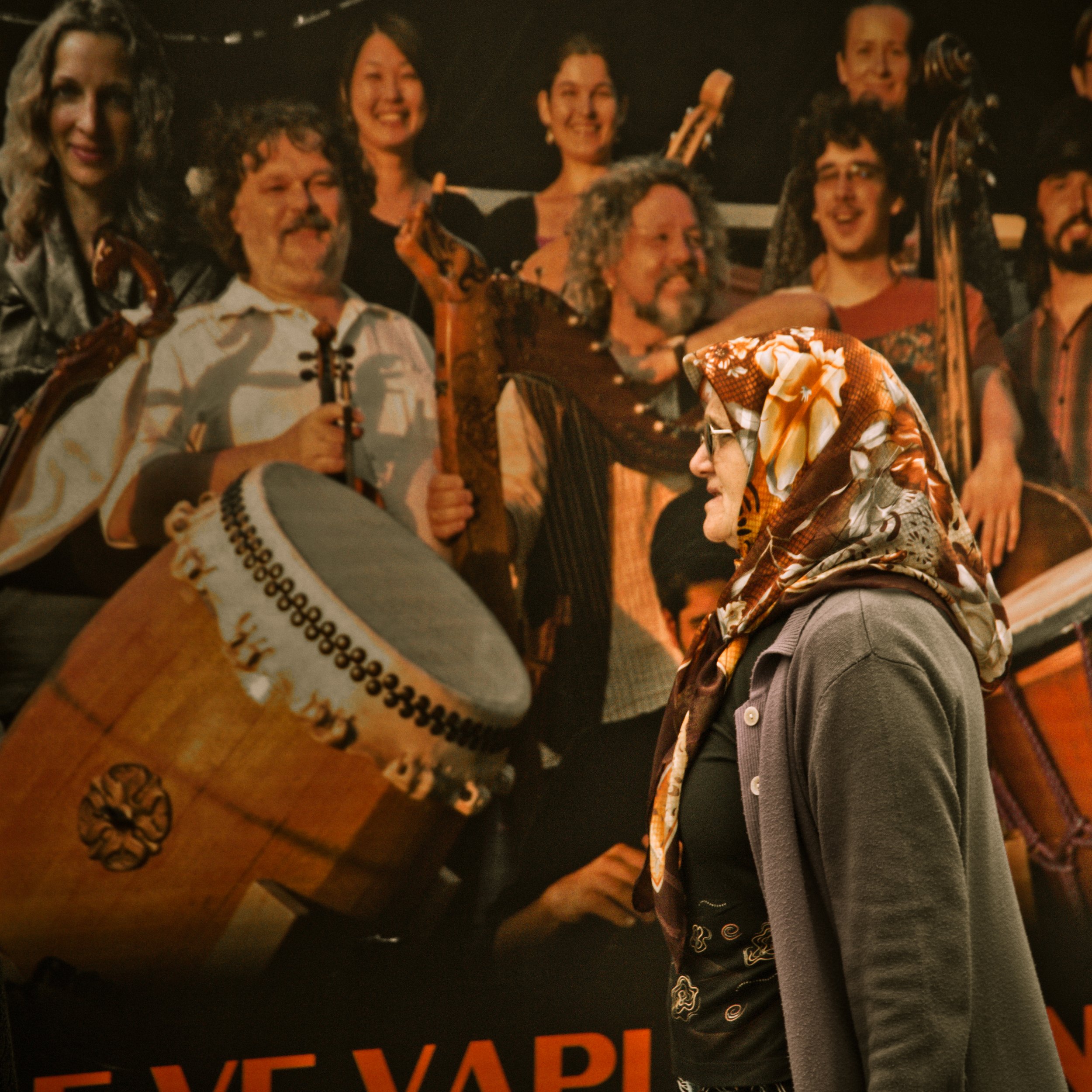 A woman wearing glasses, a patterned headscarf, and a gray jacket stands in front of a poster featuring a group of people holding musical instruments, smiling.