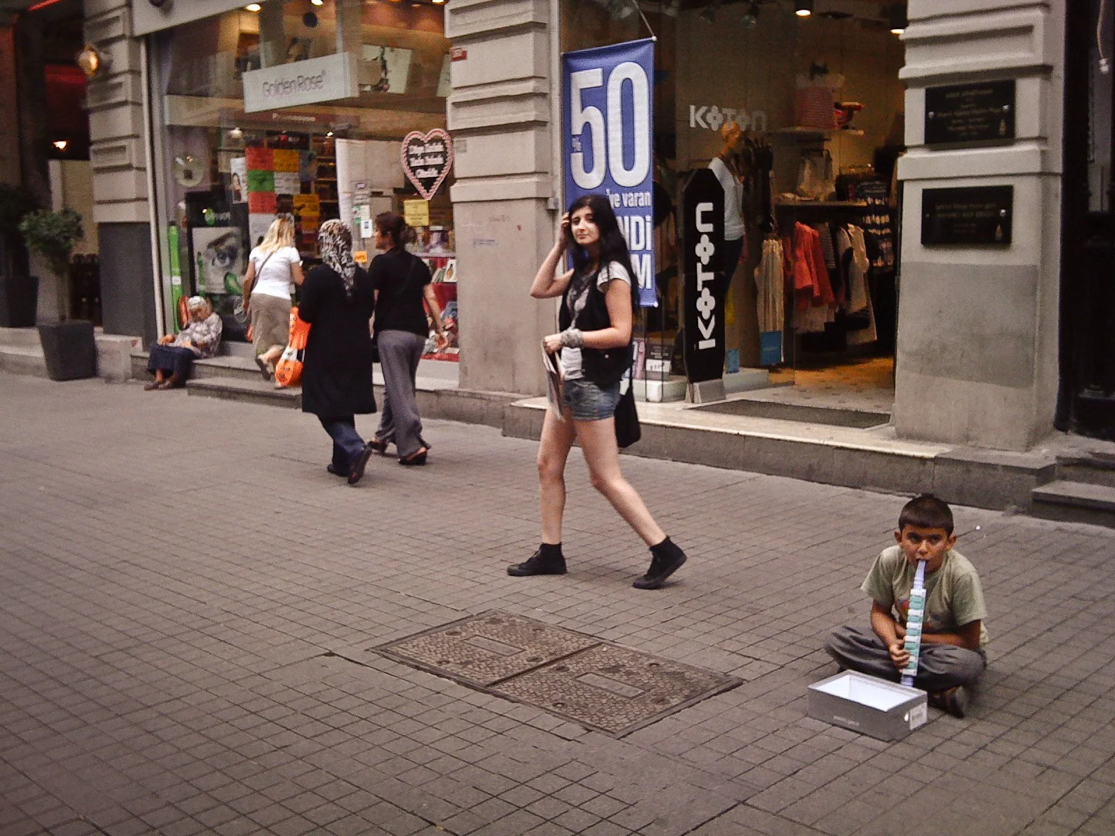 A young boy sitting on the sidewalk with a birthday cake and a candle holder in front of a shop, while a young woman walks by and other people stand near the shop entrance.